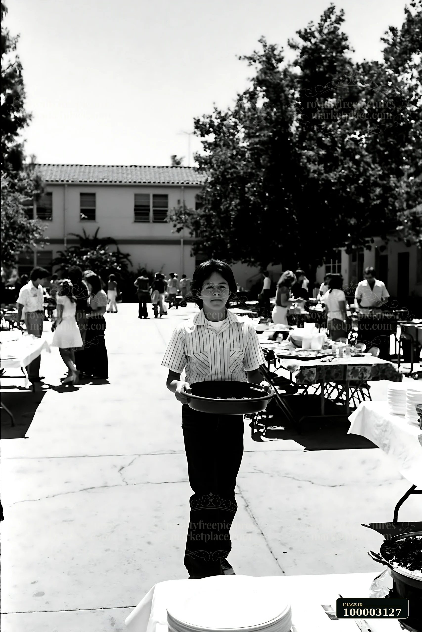 A boy holds a tray of food at a community potluck buffet held outdoors in a school yard.