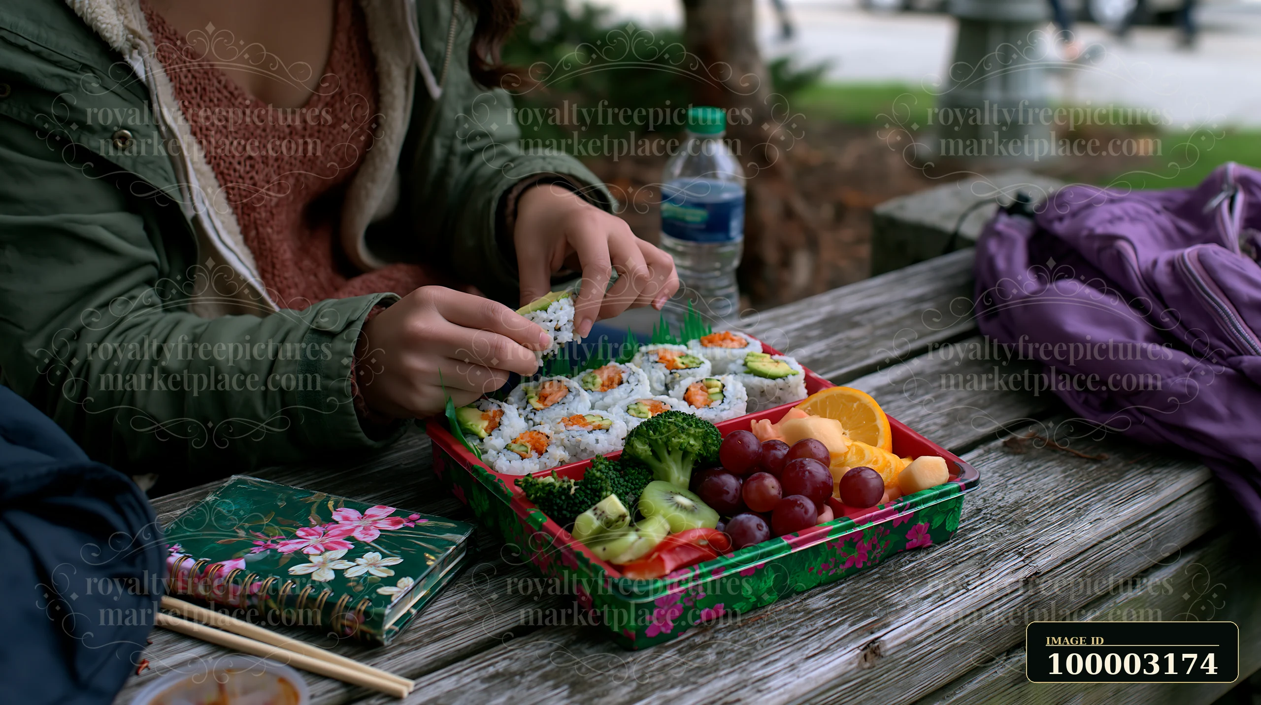 A person sits at a wooden table outdoors, preparing to enjoy a sushi lunch box with compartments filled with sushi rolls, fruit, and vegetables.