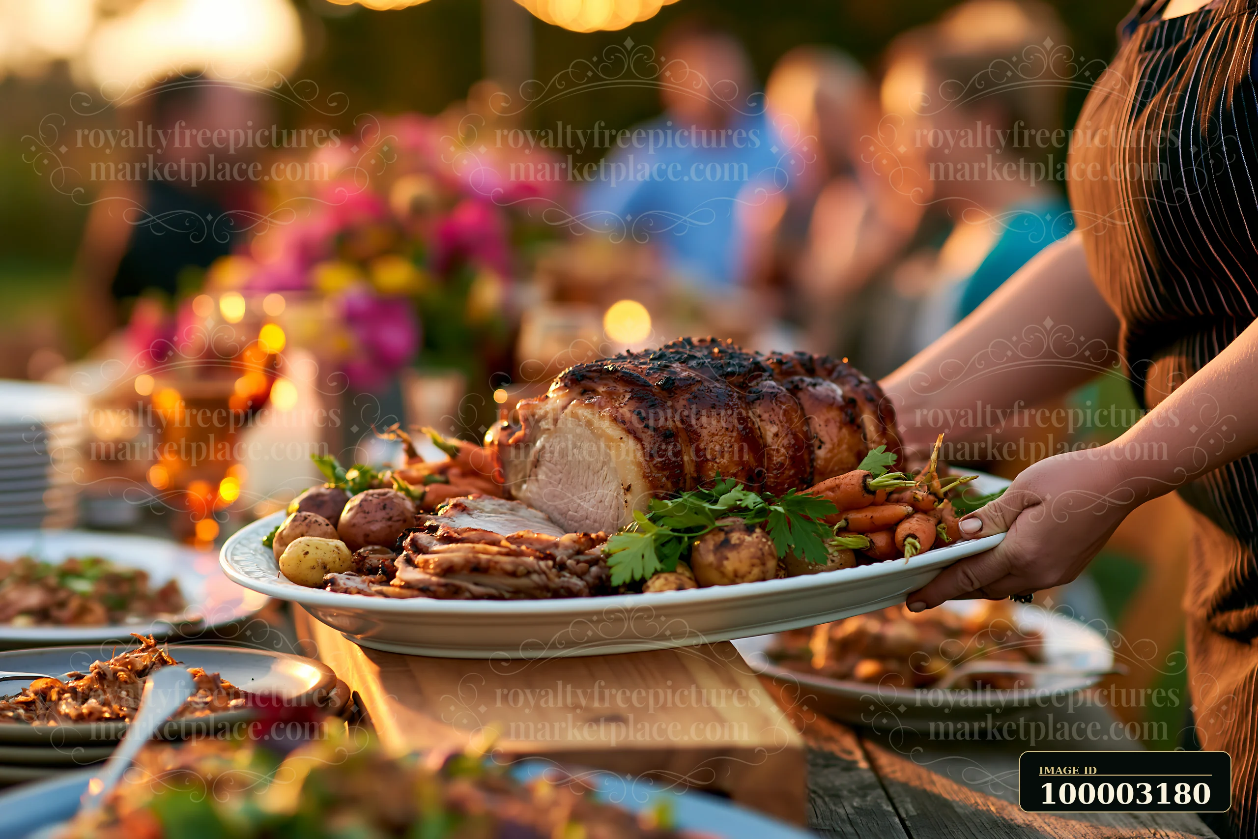 A person carries a large platter with pork roast and sides during a festive gathering at dusk.