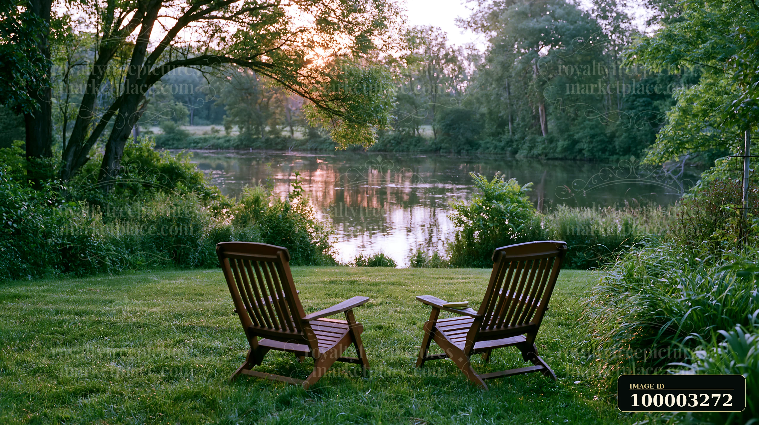 Two wooden chairs sit on a freshly mowed meadow by a river, surrounded by trees and grass.