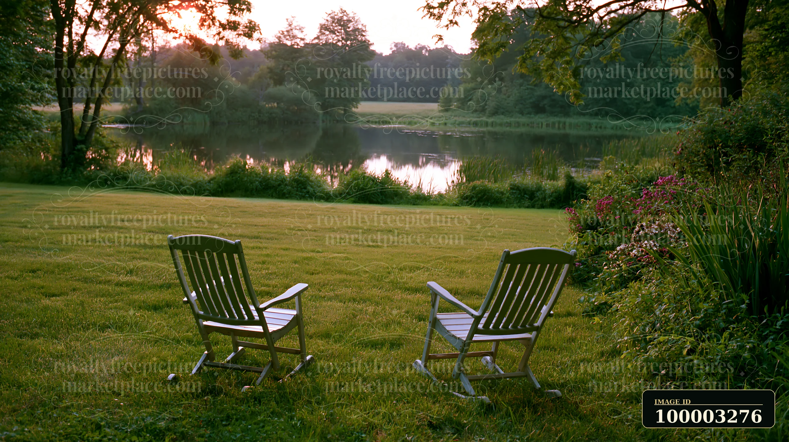 Two rocking chairs sit beside a freshly mowed meadow, facing a calm lake surrounded by trees and flowers.