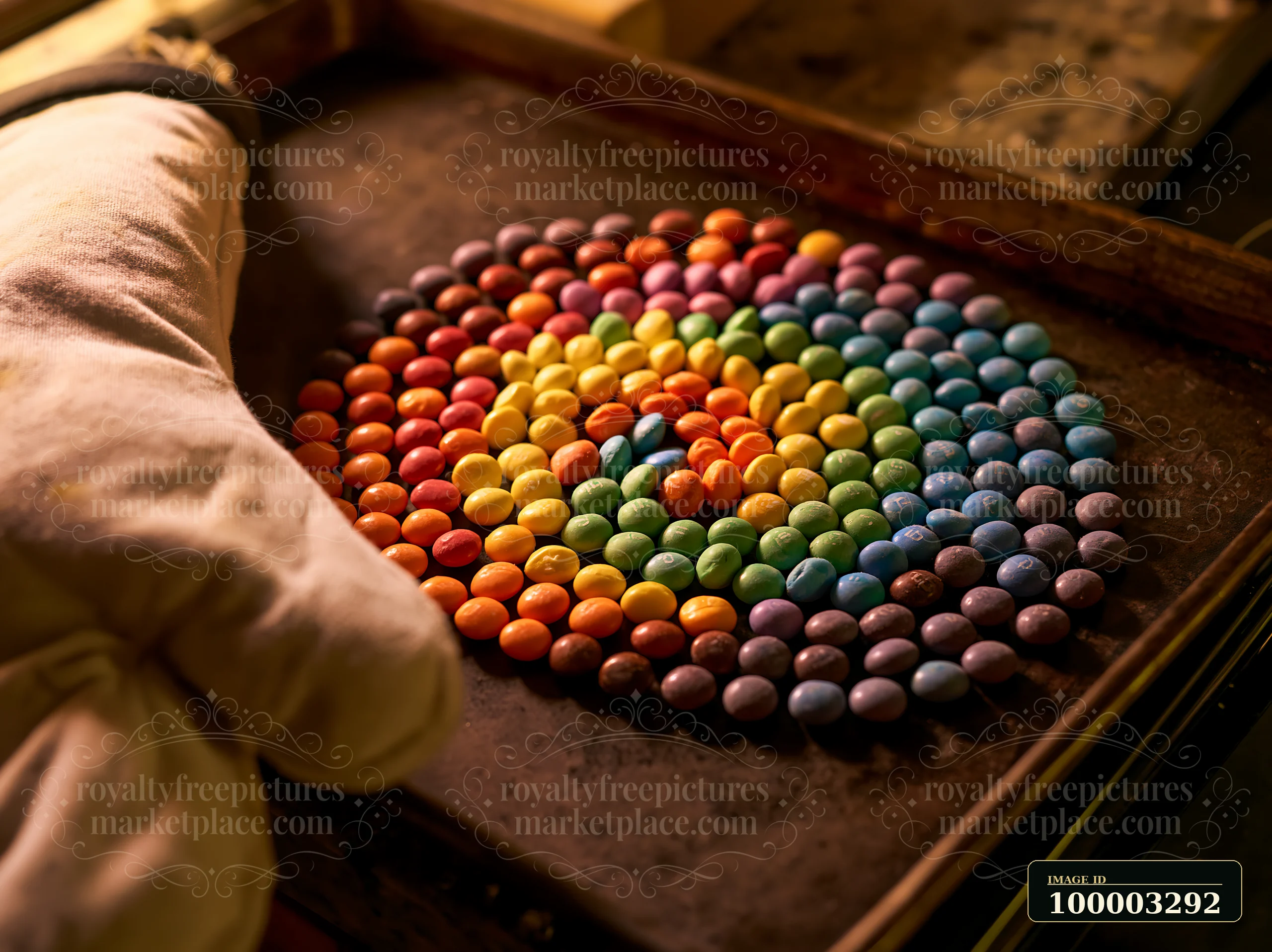 A hand in an oven mitt arranges colorful candy pieces forming spiral on a baking tray.