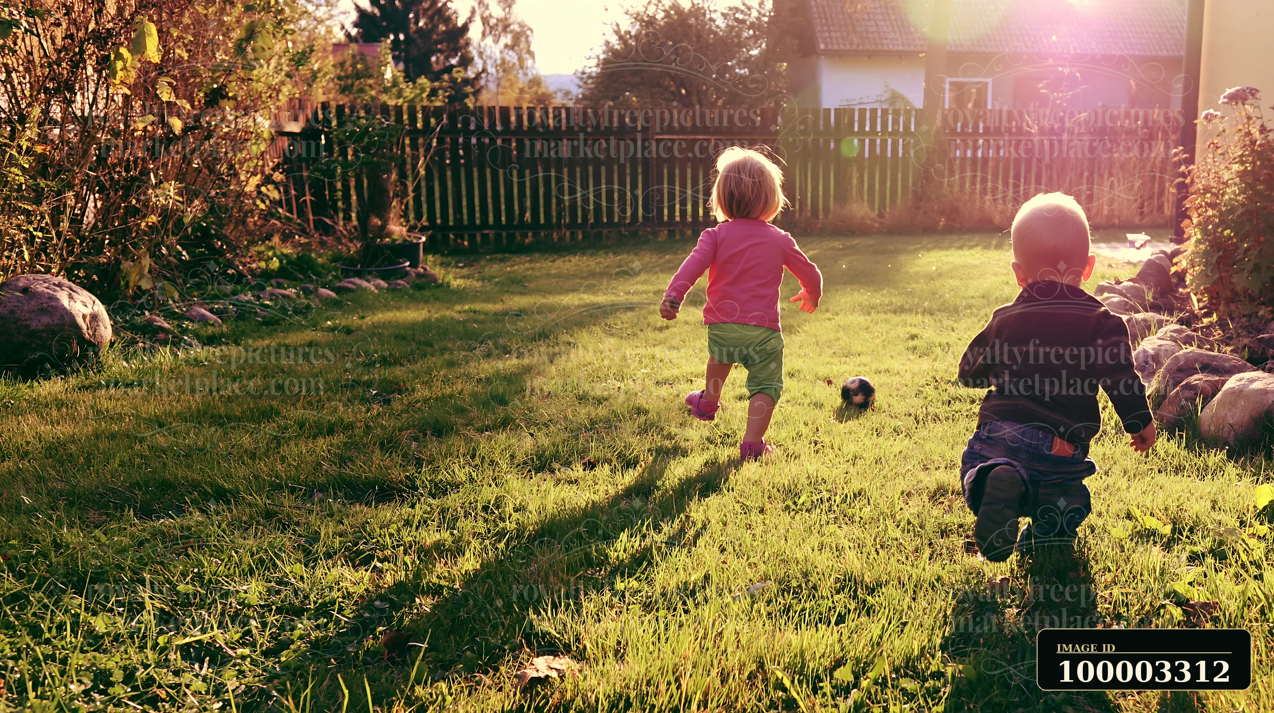 Two young children play in a farmhouse garden, running on the grass as the sun sets behind them.