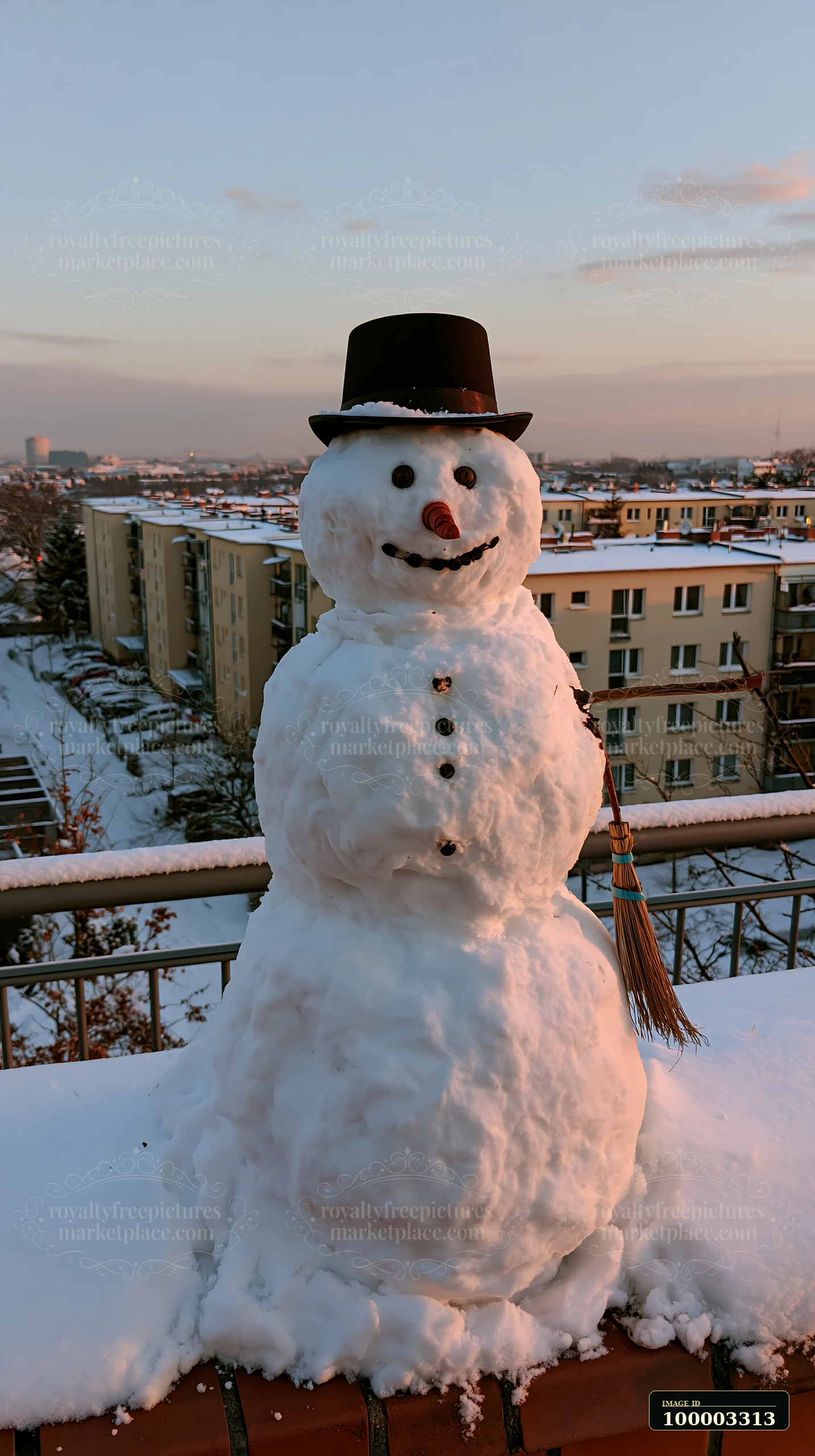 A snowman with a broomstick arm stands on a balcony with a view of apartment buildings.