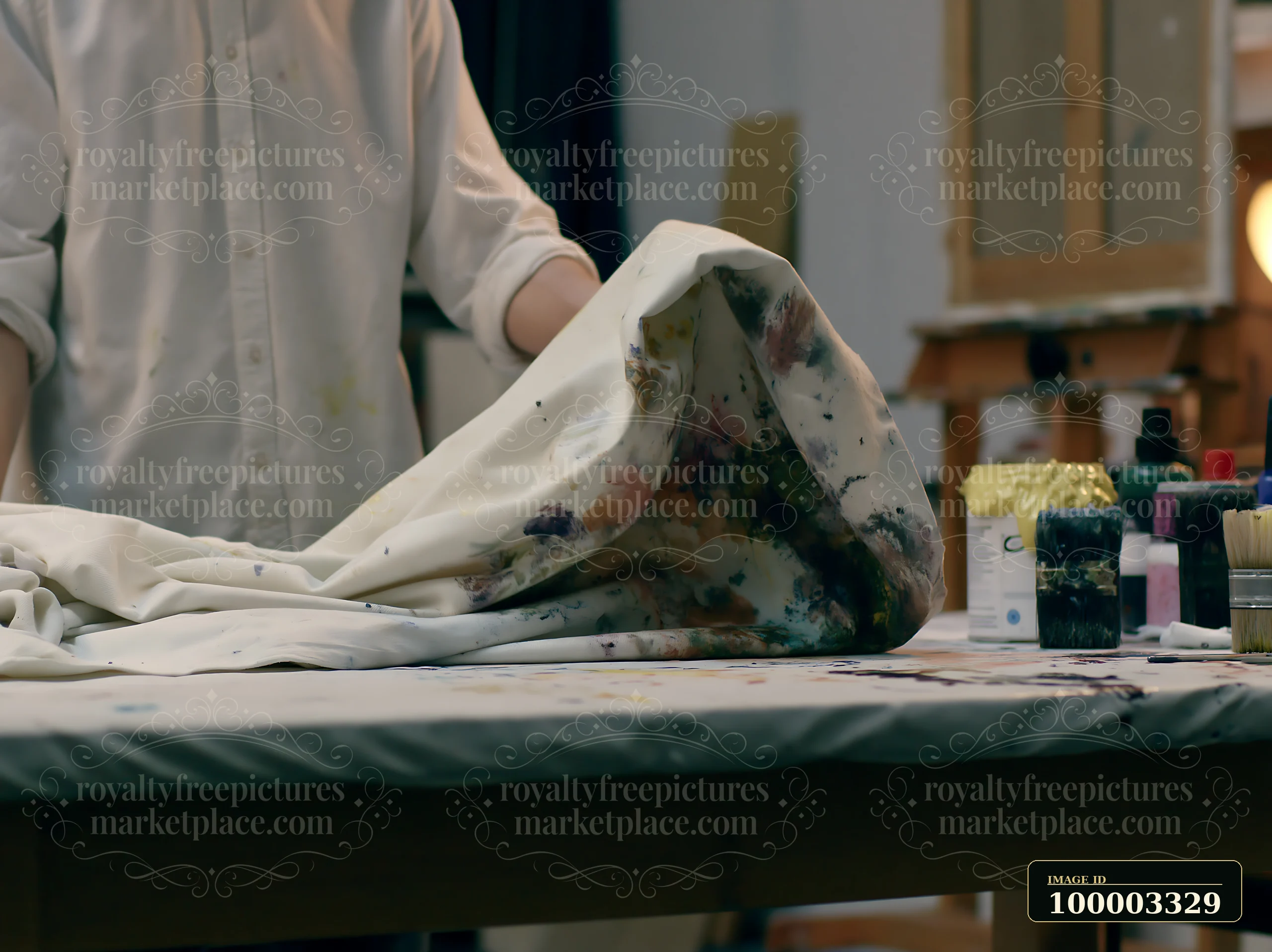 An artist works at a table covered with a splattered artist canvas textile and various painting supplies.
