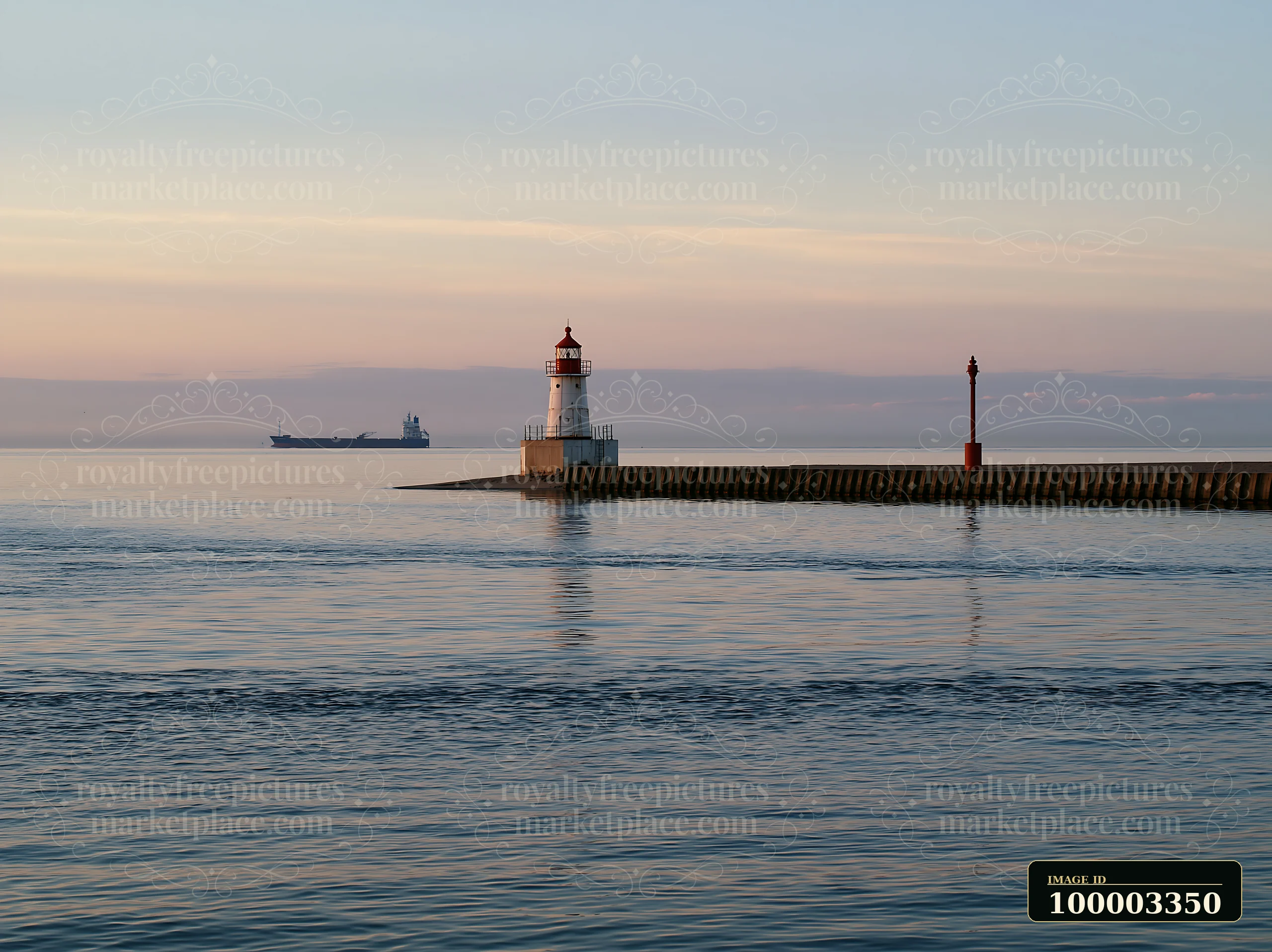 A lighthouse stands on the edge of a pier while a cargo ship can be seen in the distance across the calm water during sunset.