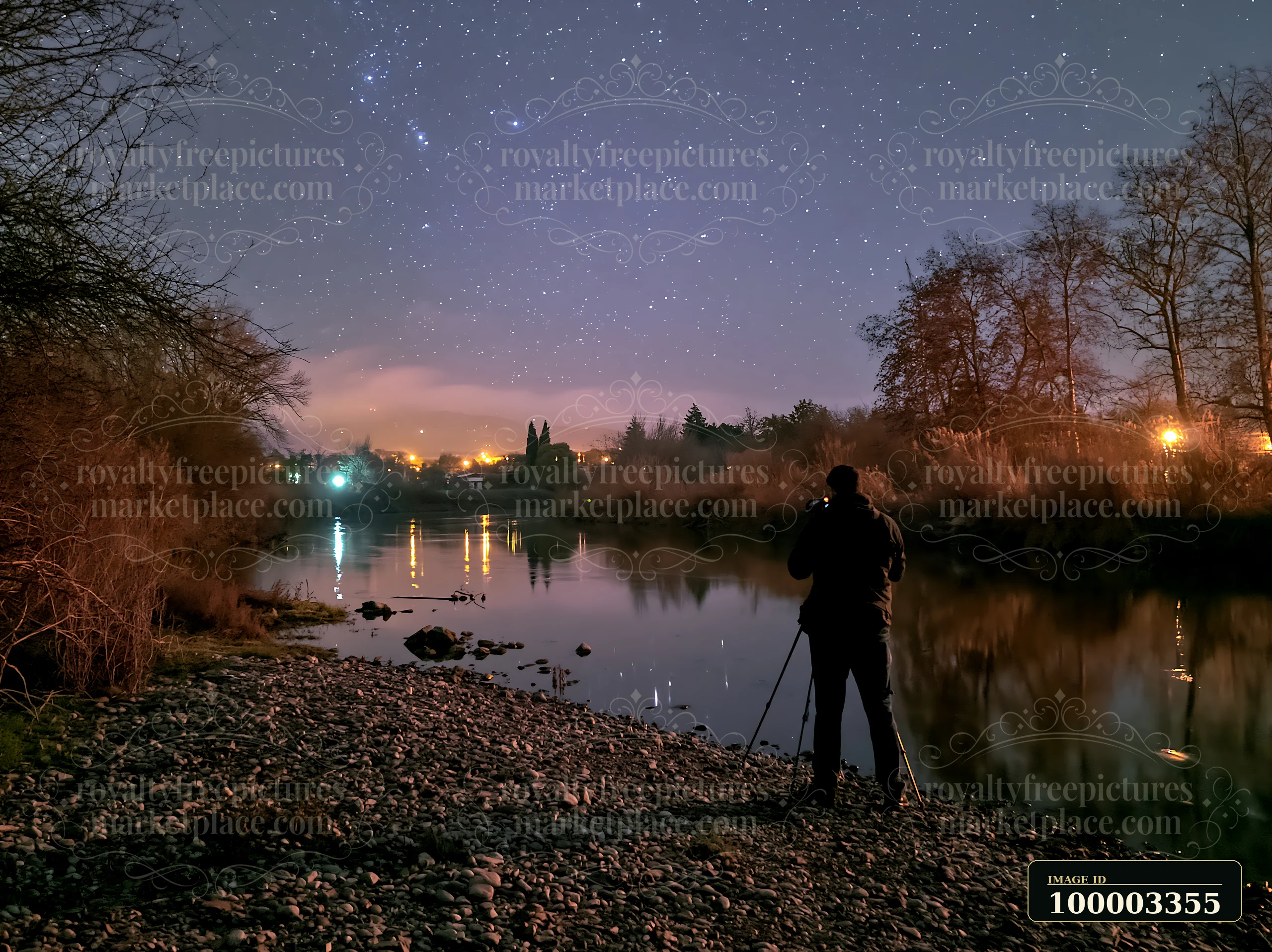 A person stands with a camera on a tripod beside a riverbank at night, capturing the starry sky.