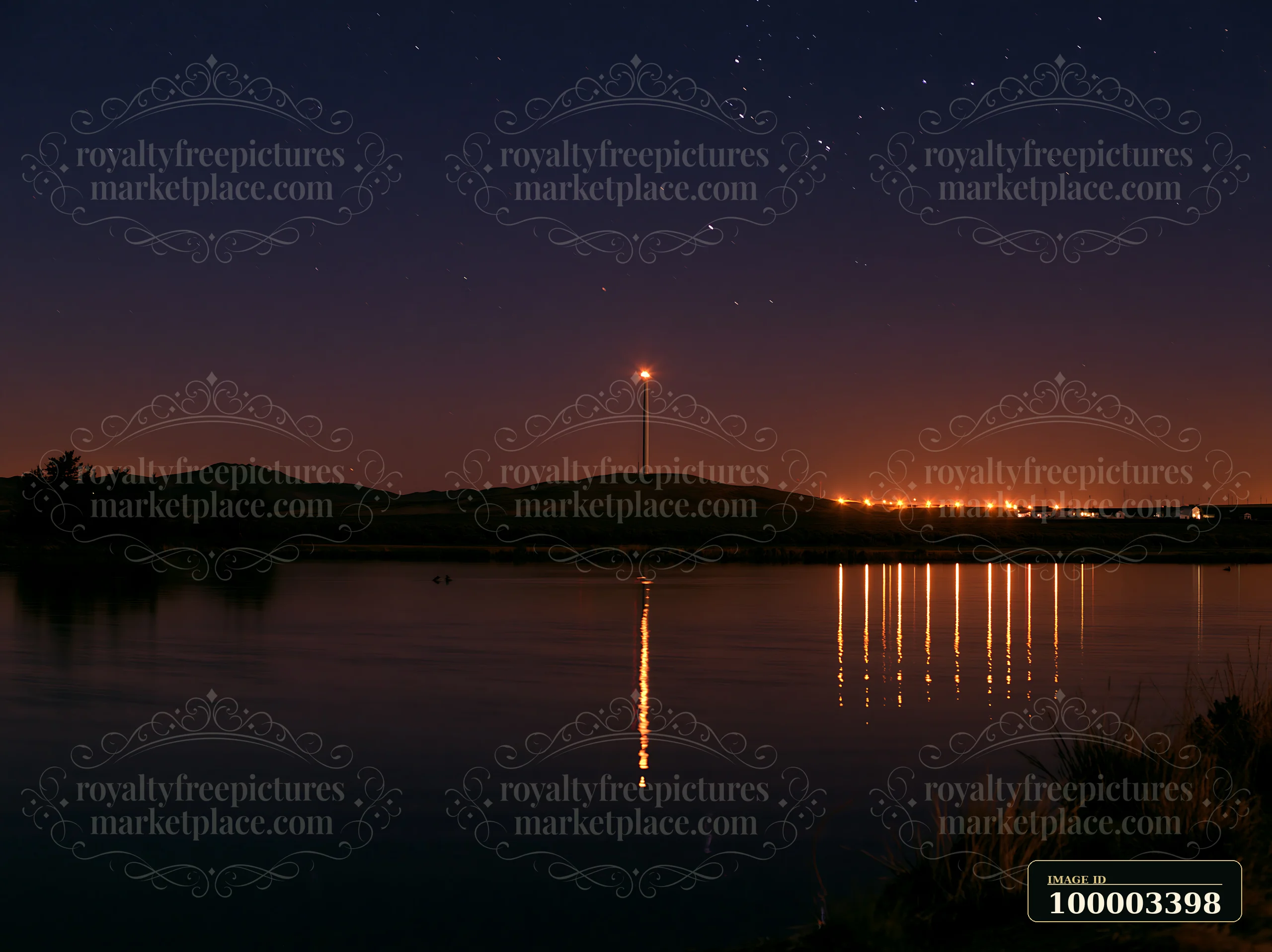 Stars fill the night sky above a rural wind turbine near a calm lake.