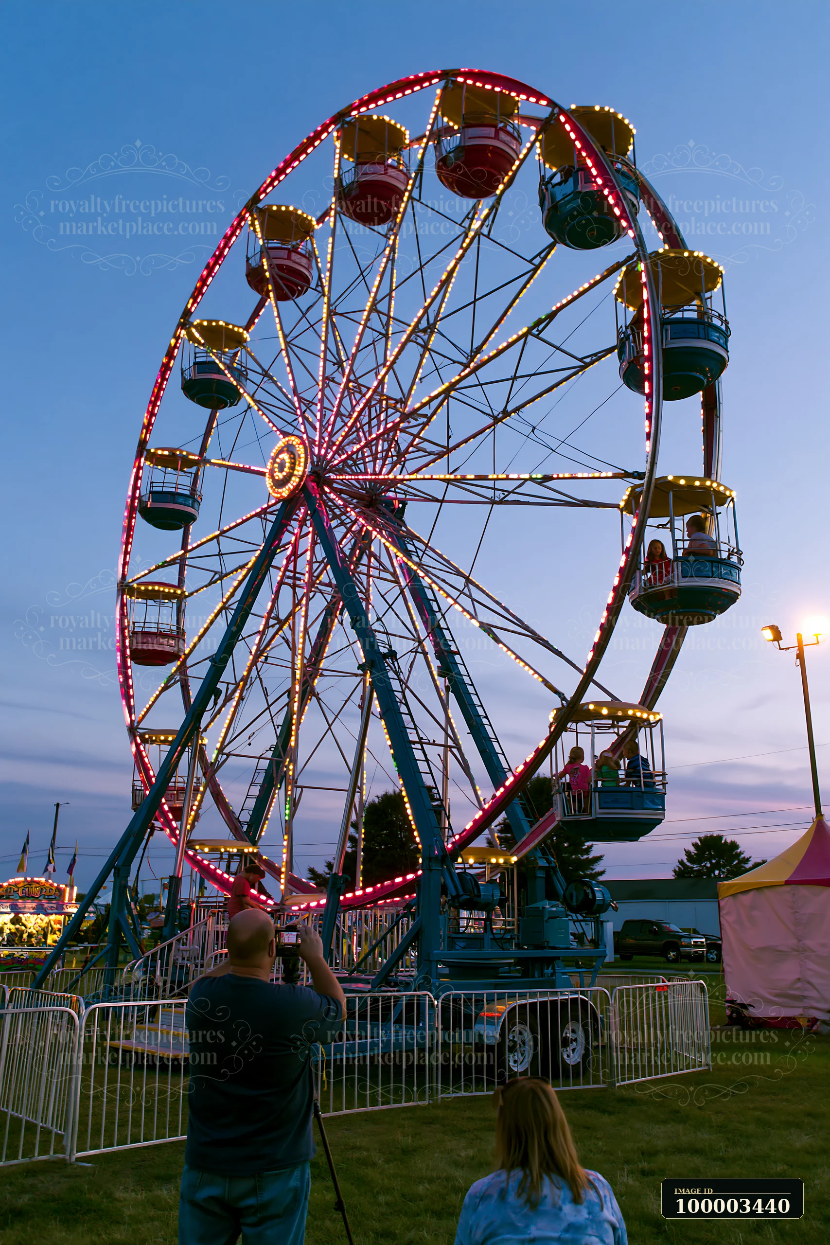 Families enjoy a fair at dusk, with parents photographing children on rides.