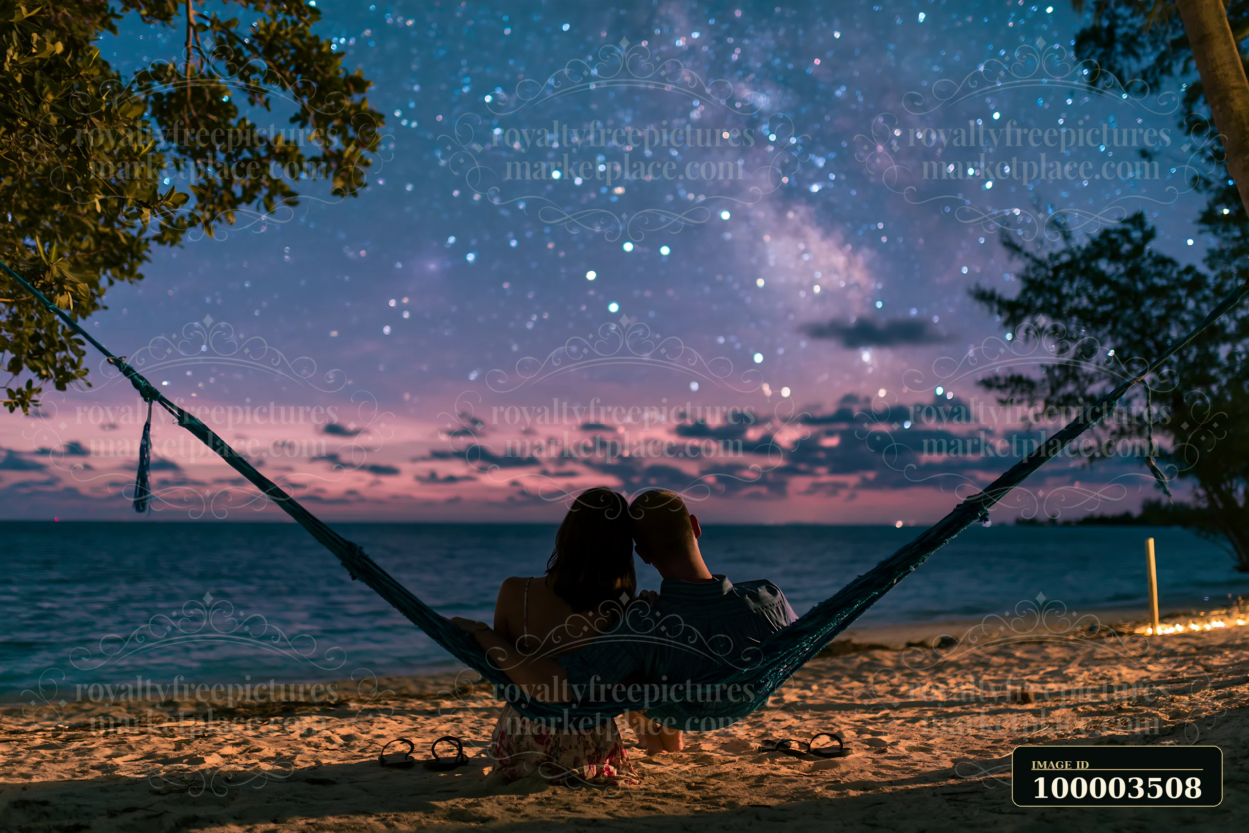 A couple sitting in a hammock under stars views the night sky near the beach.