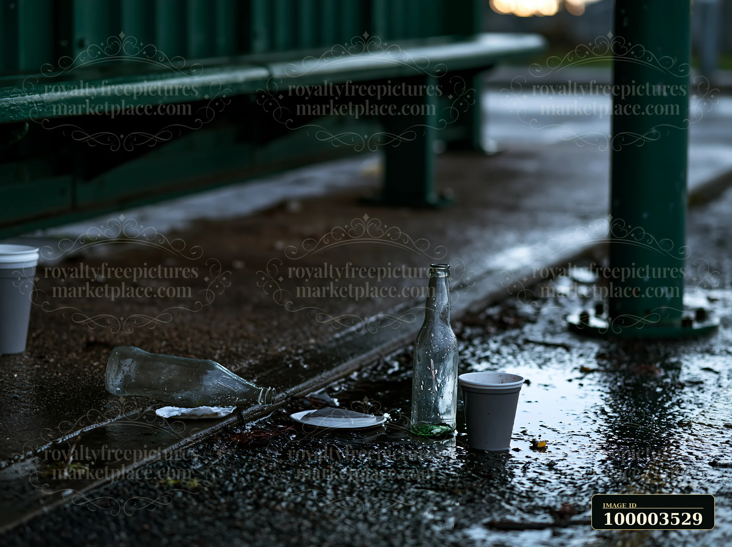 A clear bottle sits next to paper cups and a plate on a damp ground in an outdoor setting, showing litter left behind in a public space after an event or gather