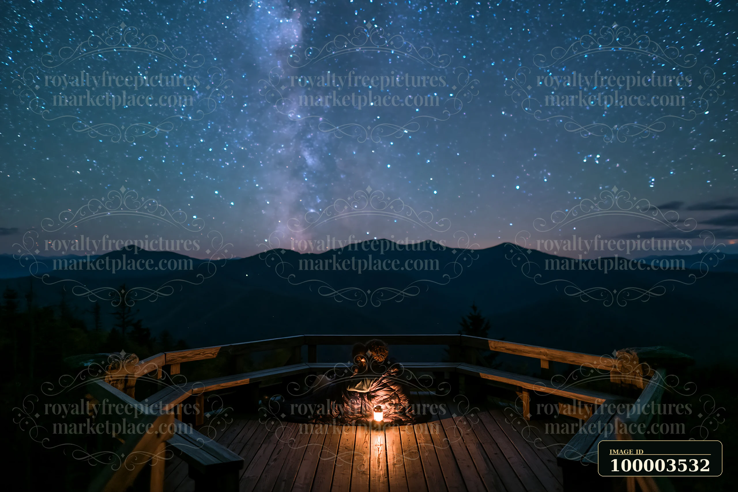 A couple sharing a sleeping bag beneath stars sits on a wooden deck, enjoying the view of mountains and the Milky Way overhead while a warm light glows from a n