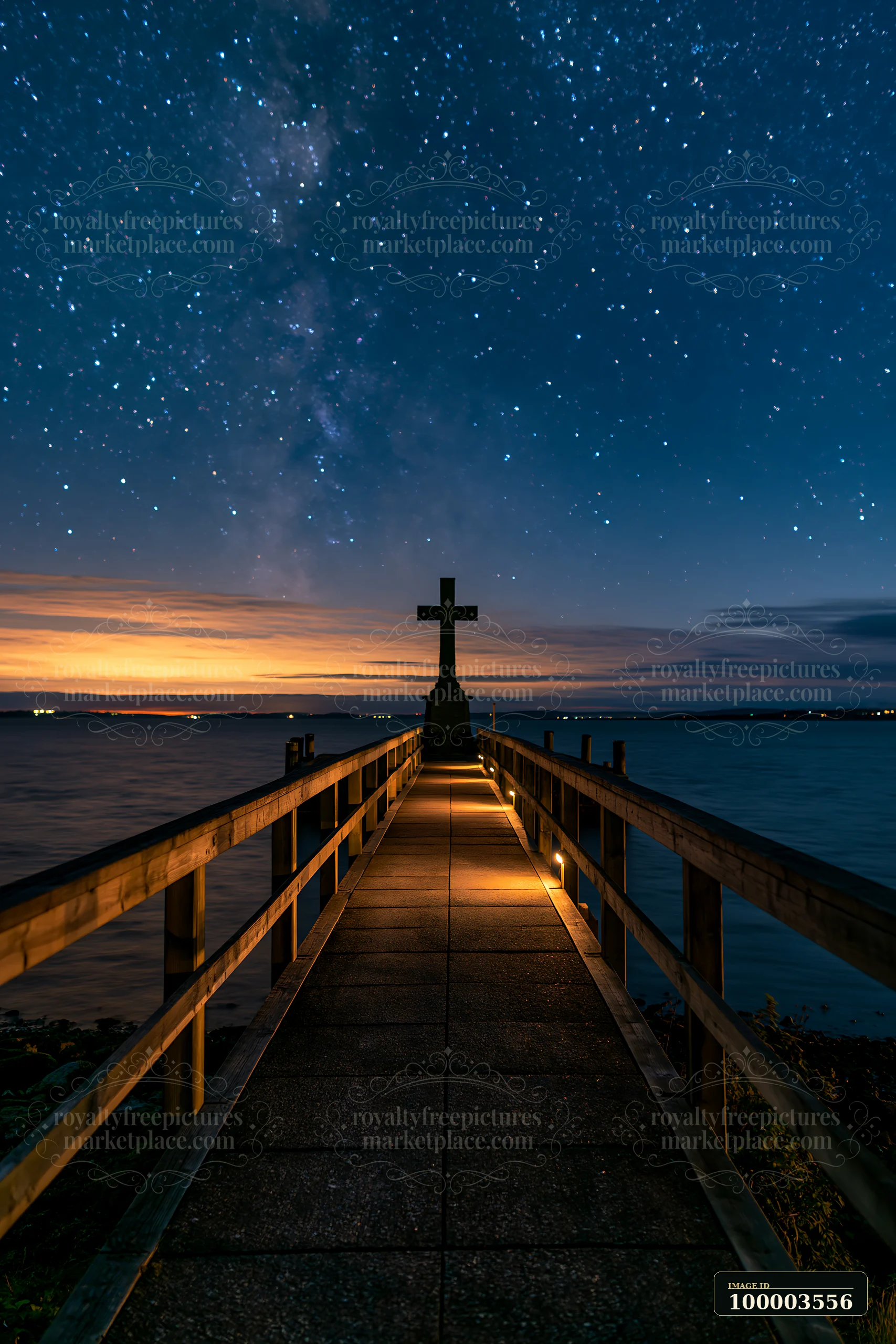 A war memorial cross stands on a wooden pathway leading to the water, illuminated by low lights, with a starry sky and colorful sunset in the backdrop.