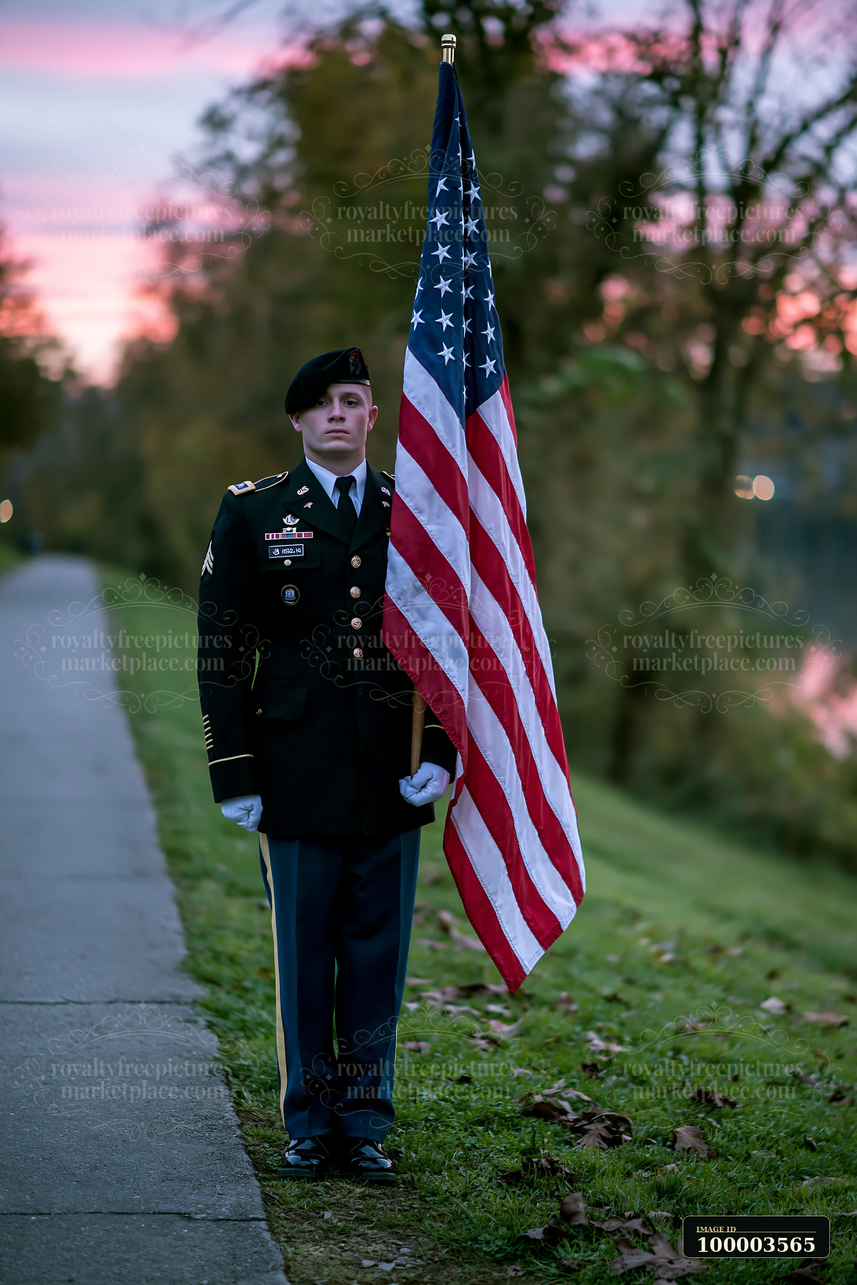 A soldier stands at attention while holding the American flag near a path beside a river at sunset.