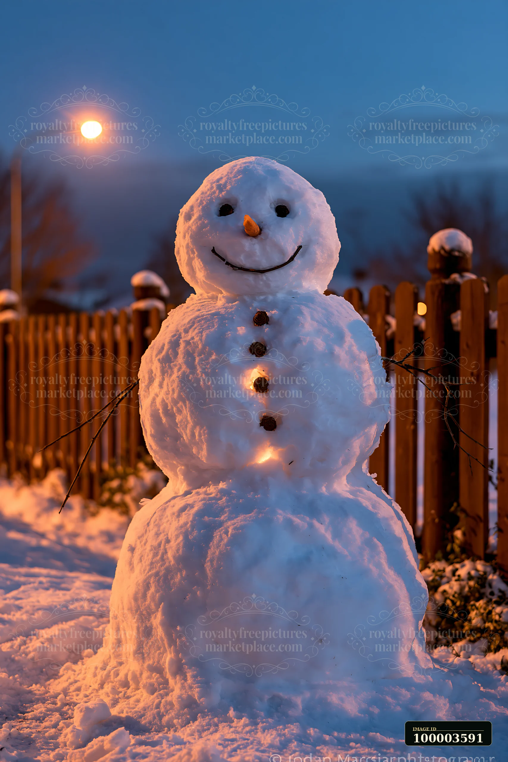 A cheerful snowman stands beside a wooden fence in the evening light.