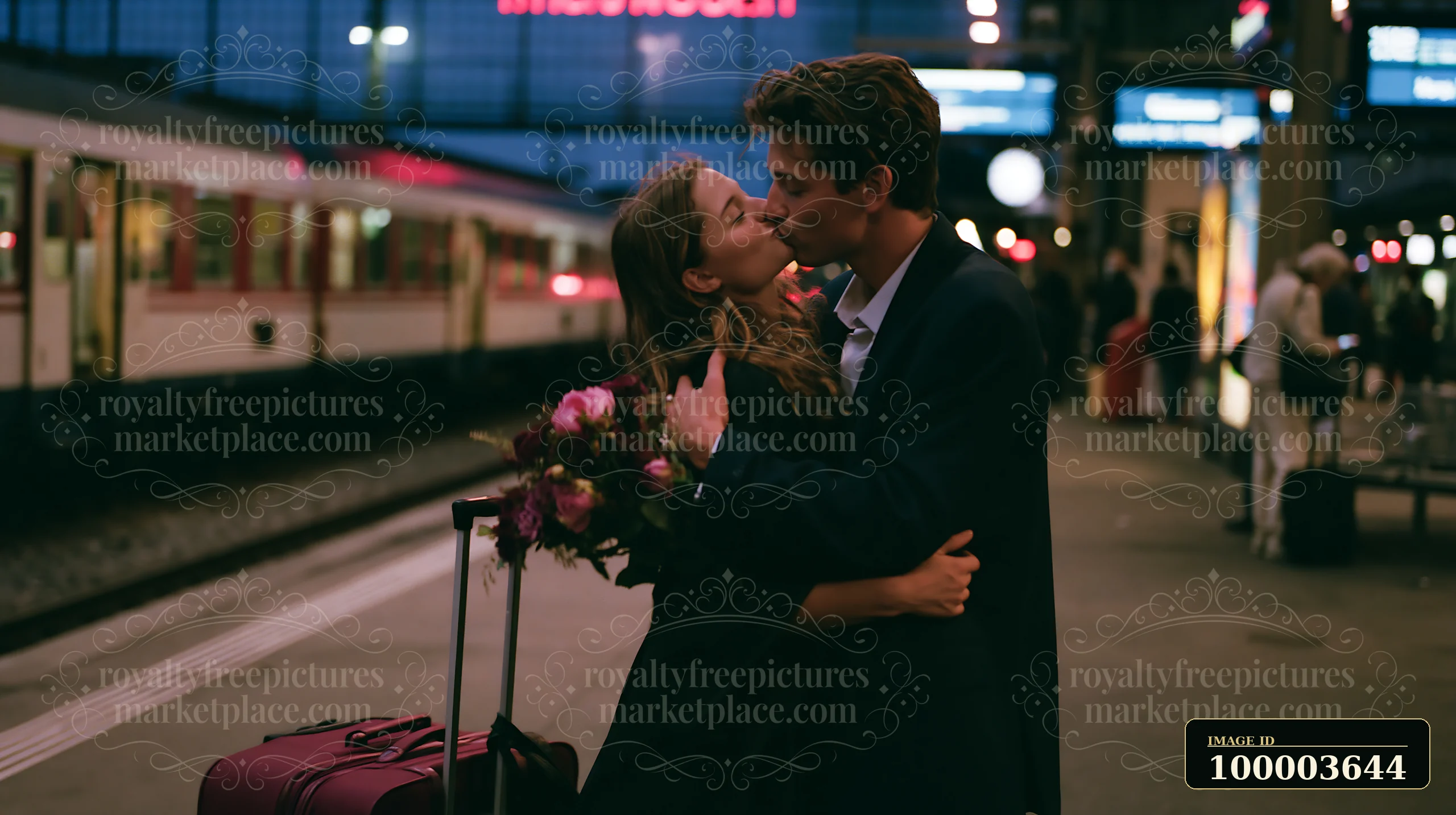 A couple kisses at a train station during their graduation celebration while holding flowers and standing next to a suitcase.
