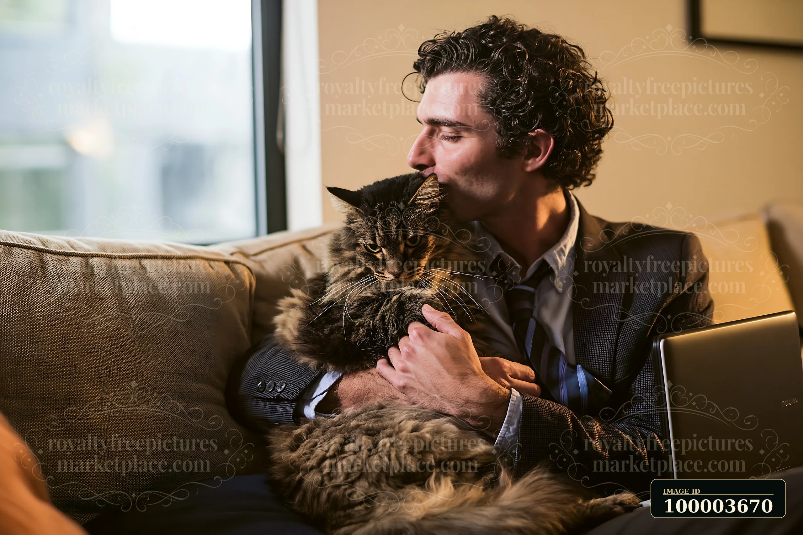 Man kisses a long-haired cat while sitting on a sofa beside an open laptop.