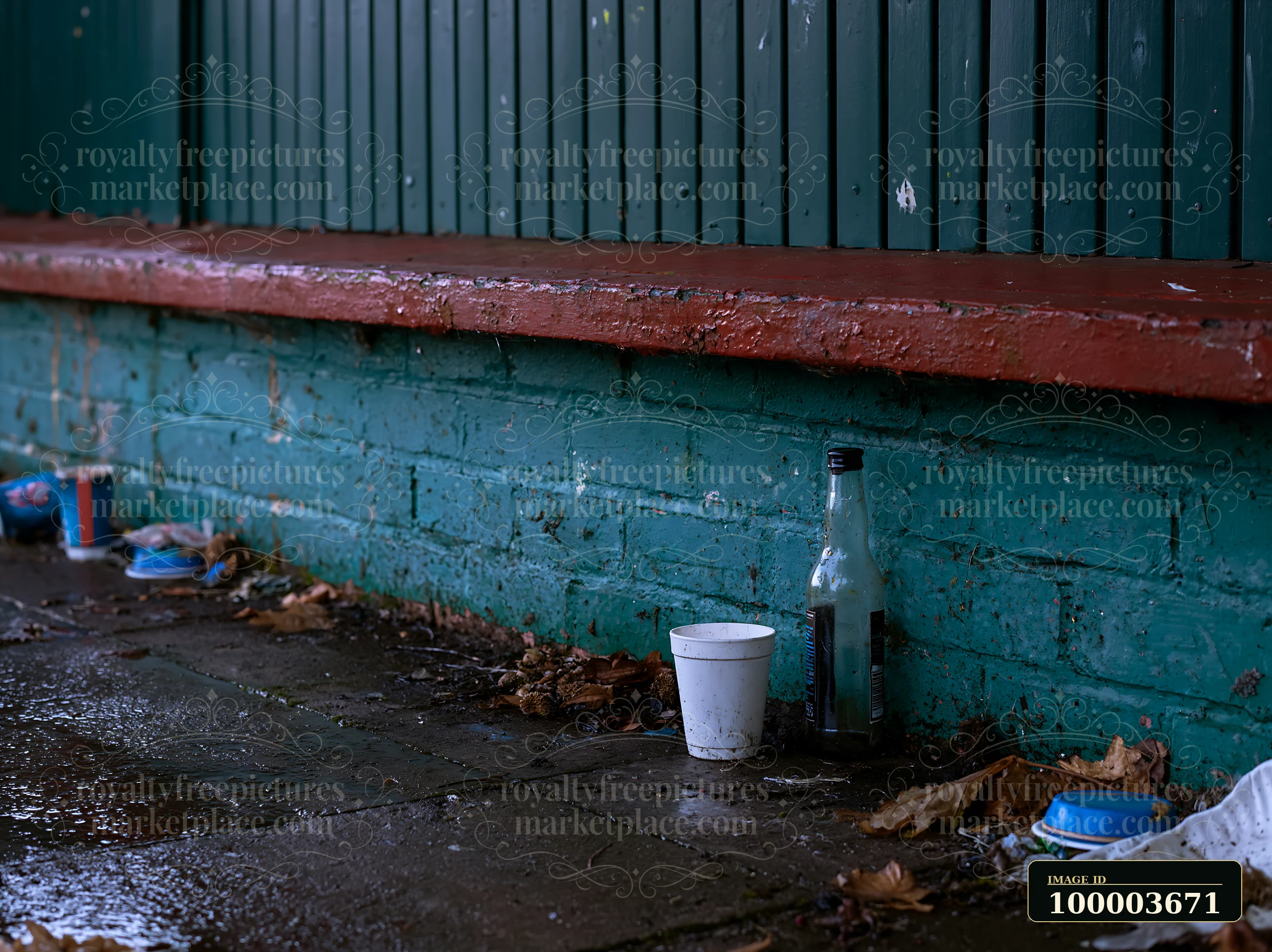 Empty bottle and discarded cups rest on wet pavement beside a weathered teal brick wall.