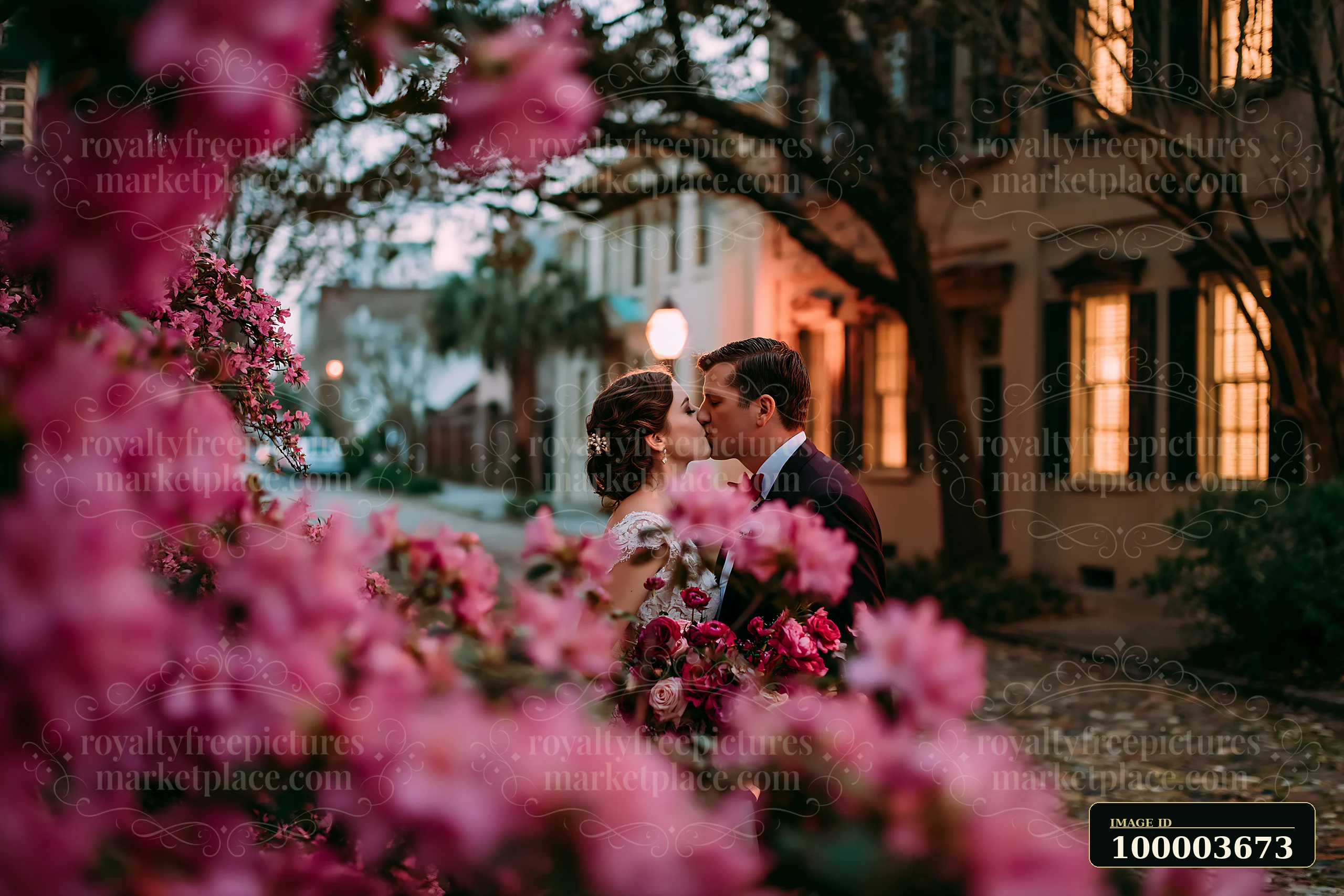 Newlyweds share a kiss outdoors in a charming street lined with blooming flowers and historic buildings in the evening light.