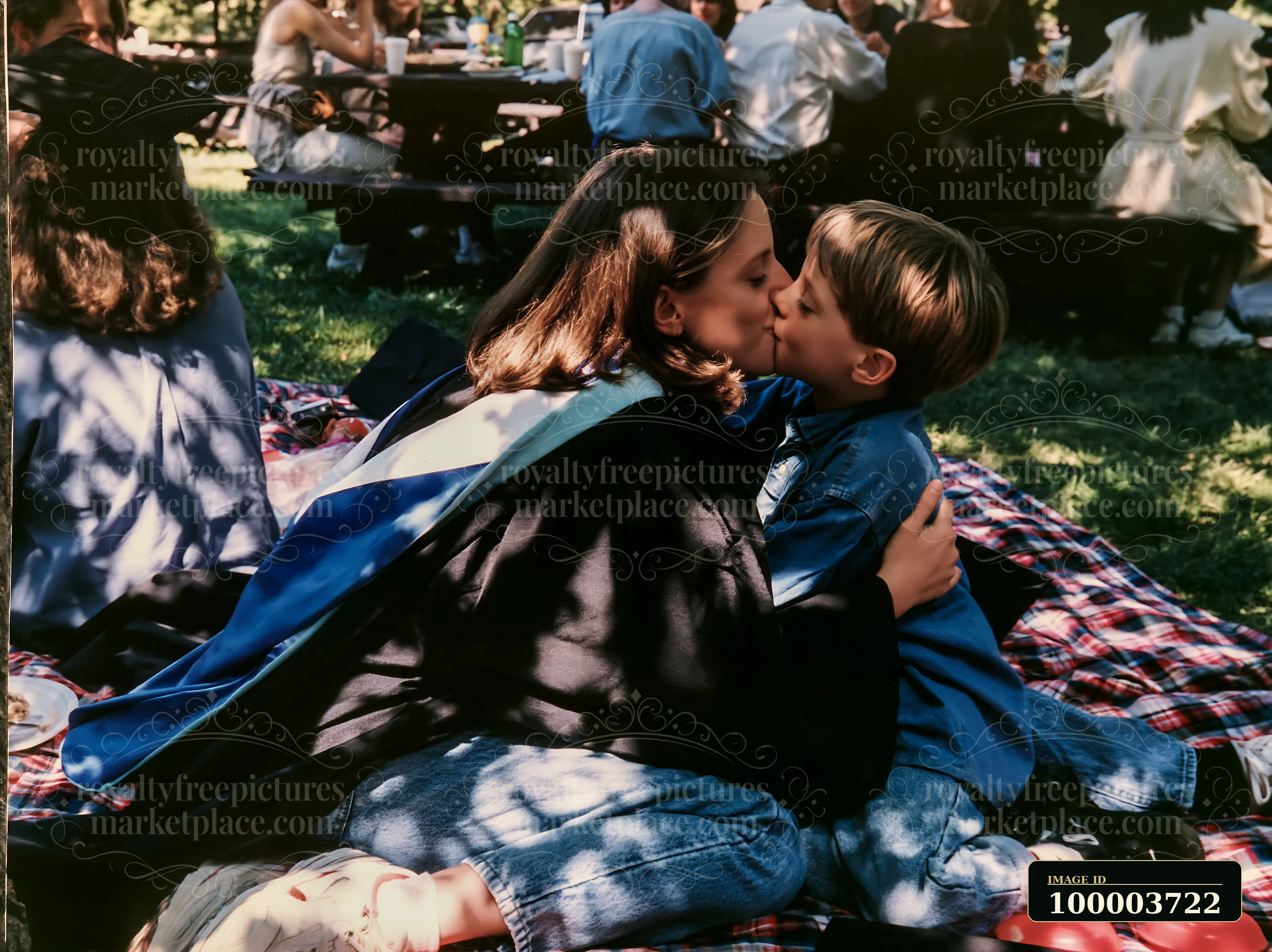 Graduate in cap and gown kisses a young boy on a picnic blanket at an outdoor celebration.