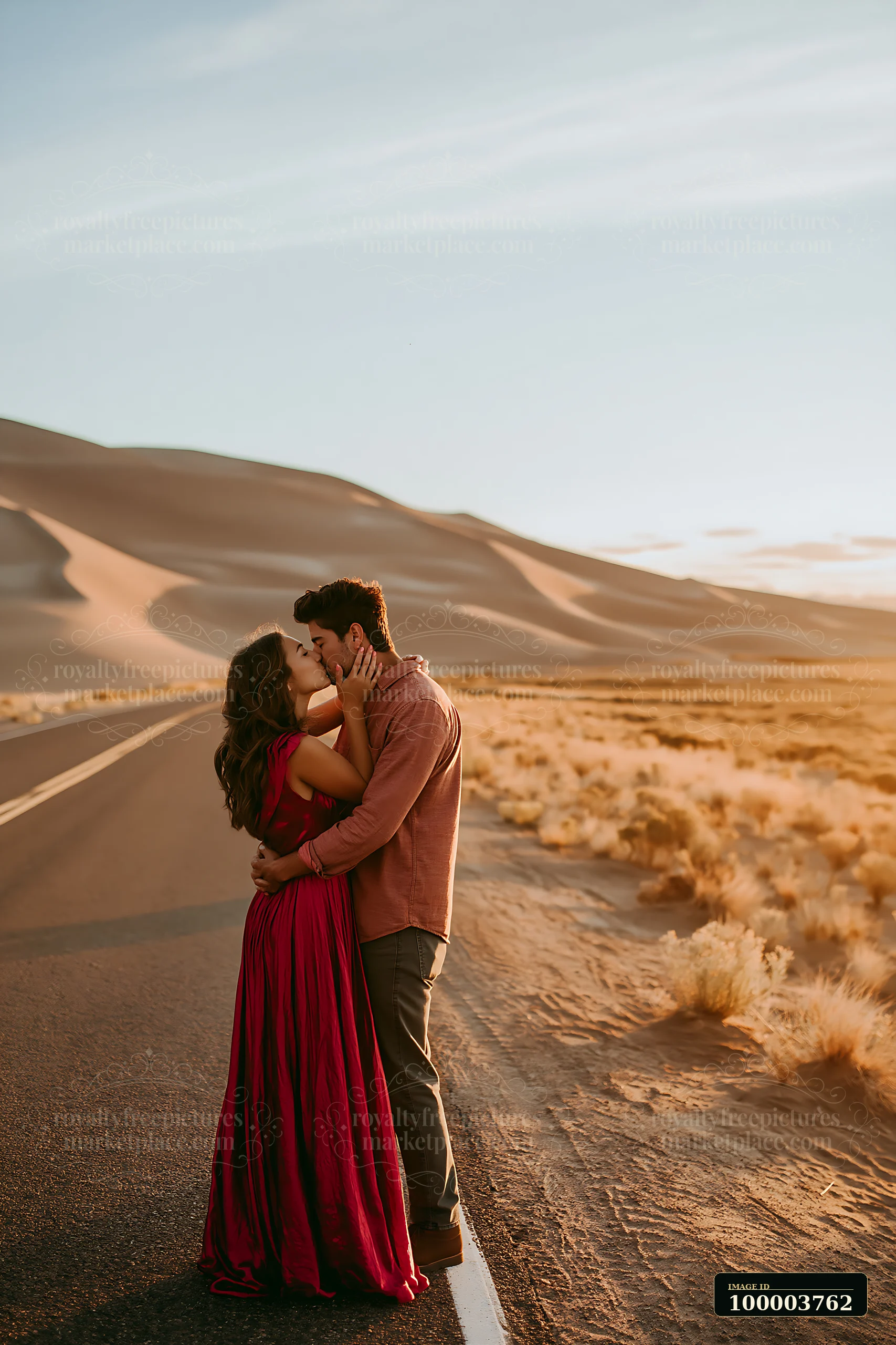 A couple is kissing on an open road surrounded by desert scenery and sand dunes during sunset.