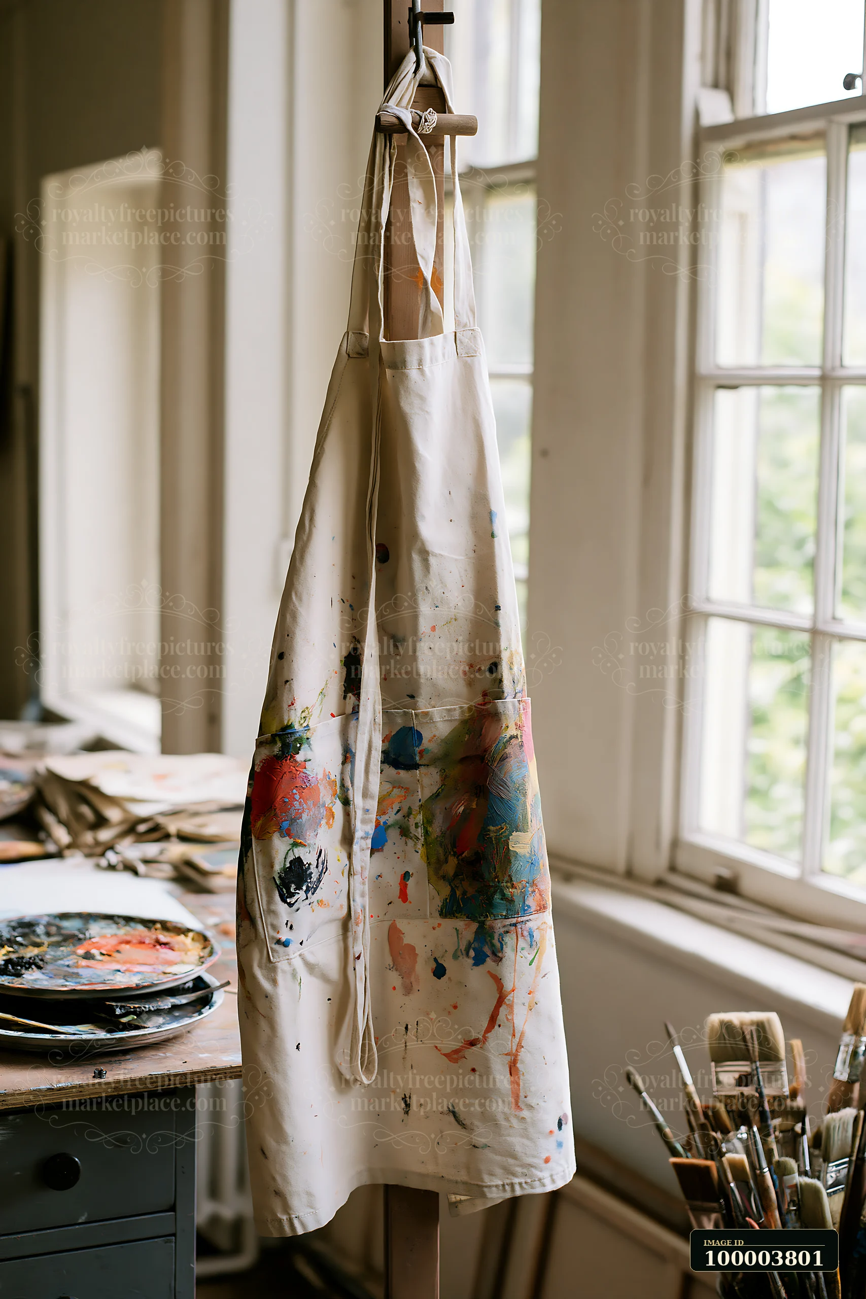 An artist apron hangs from a hook in a well-lit studio, displaying vibrant paint splatters.