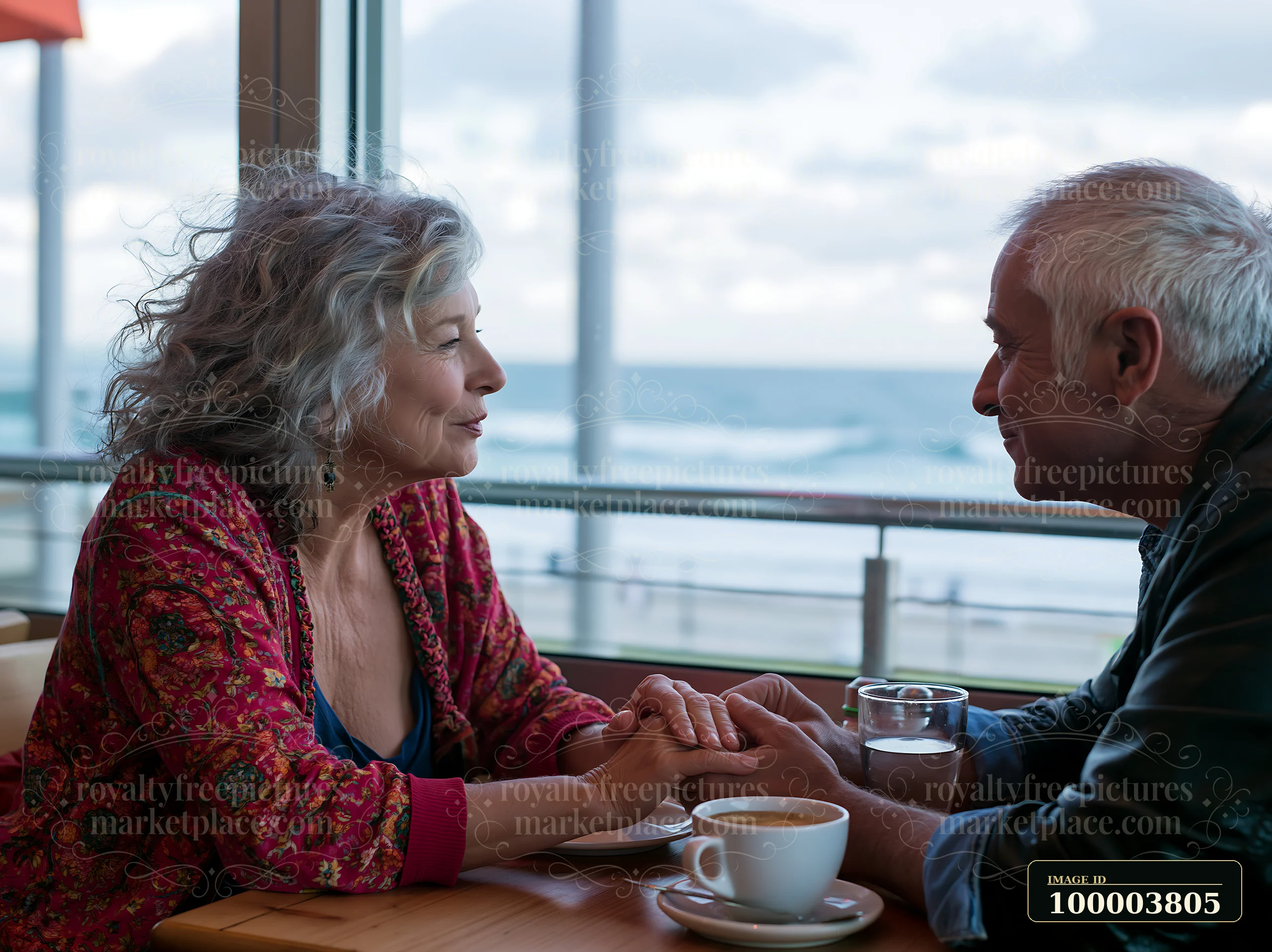 A couple sits across from each other in a cafe, holding hands and smiling, while overlooking the ocean.
