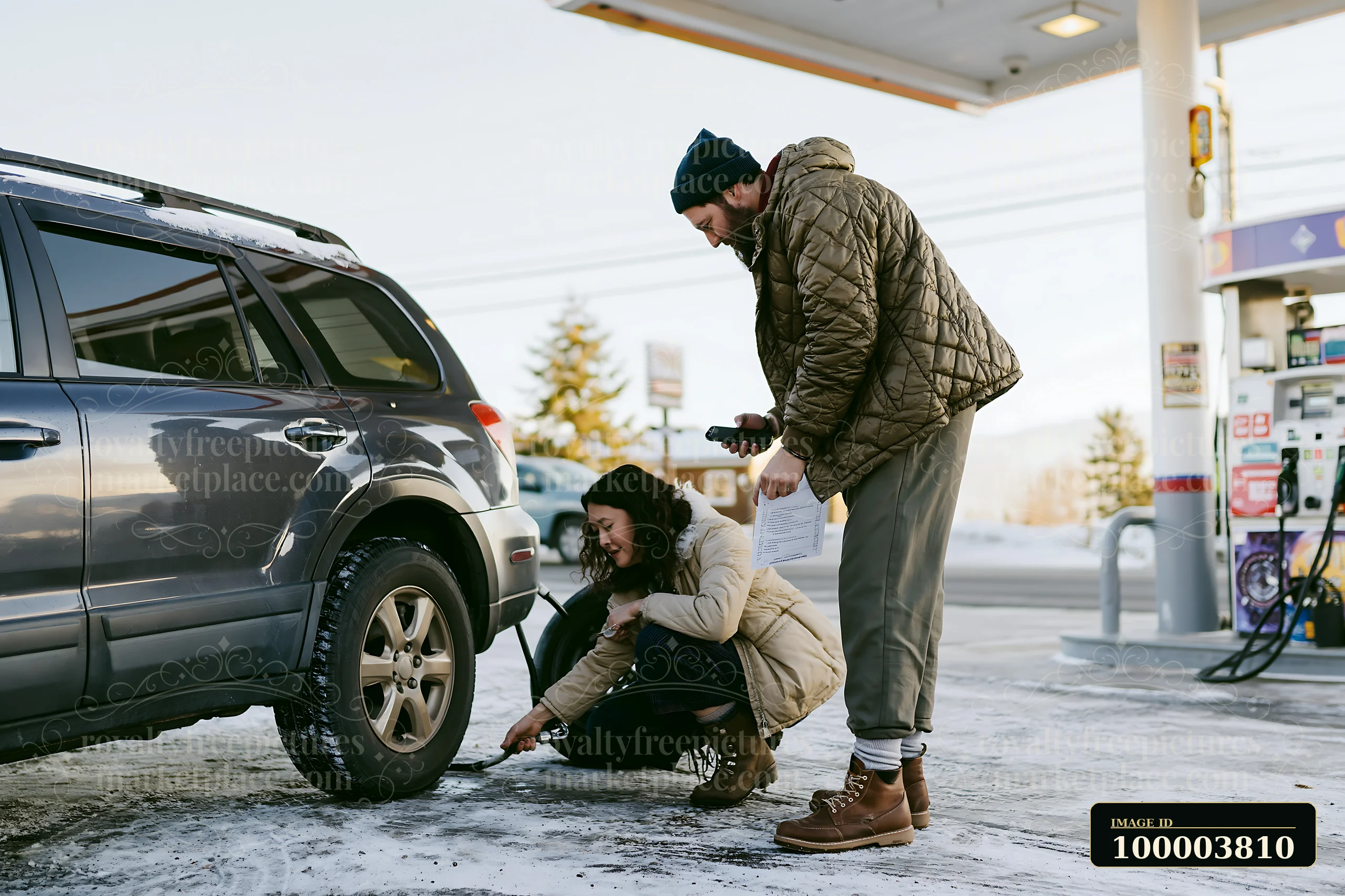 Couple prepares an SUV tire at a snowy gas station for a winter road trip.