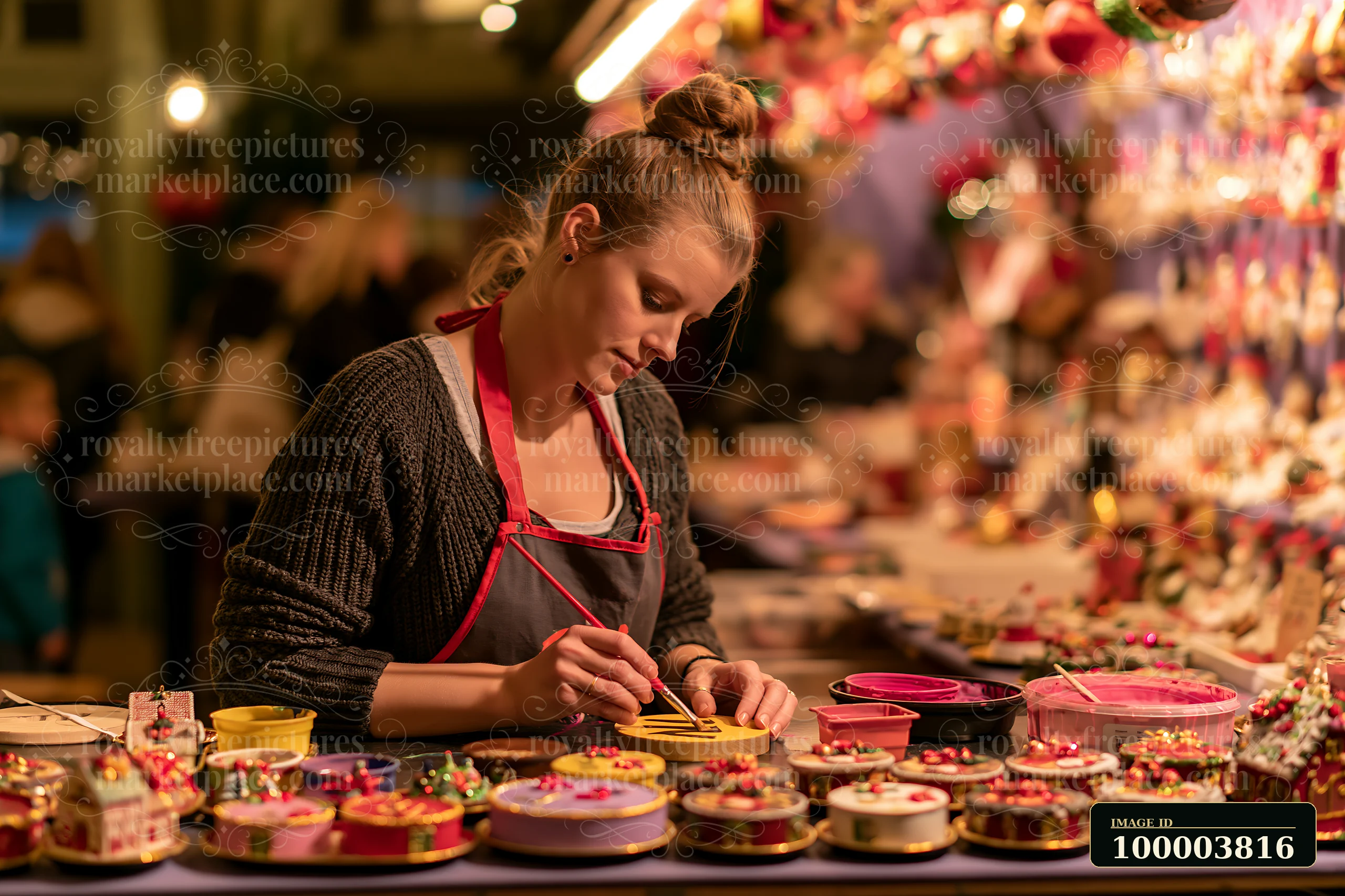 A craftsperson paints handmade ornaments at a Christmas market stall during the evening.
