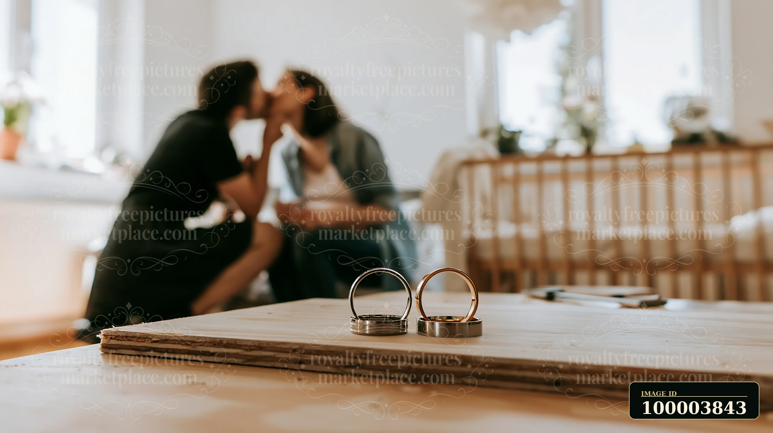 A couple kissing in a softly lit room while wedding rings rest on a wooden surface.