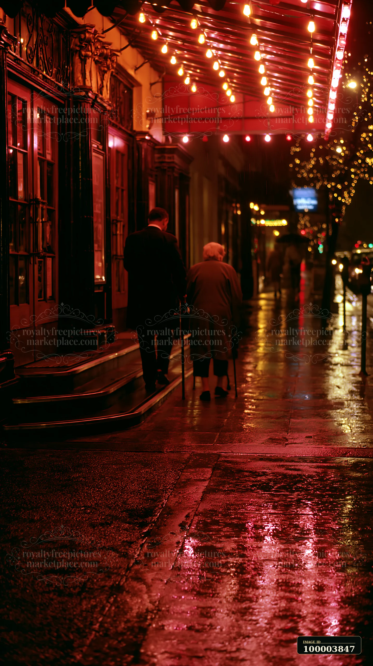 Two people walk beside a decorated storefront as Christmas lights reflect on wet pavement at night.