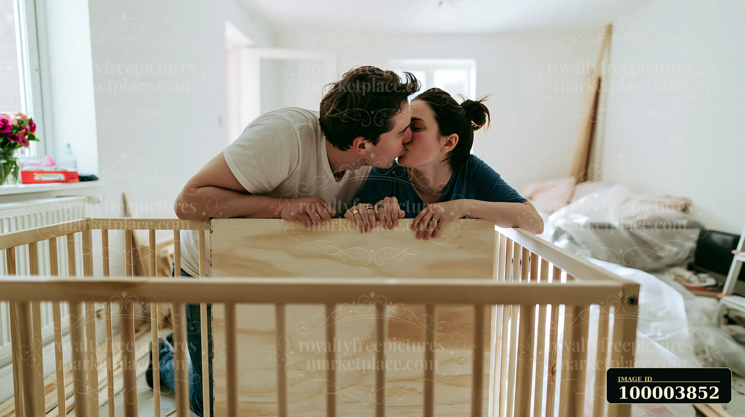 Couple kisses beside wedding rings while leaning over a wooden crib in a bright bedroom.