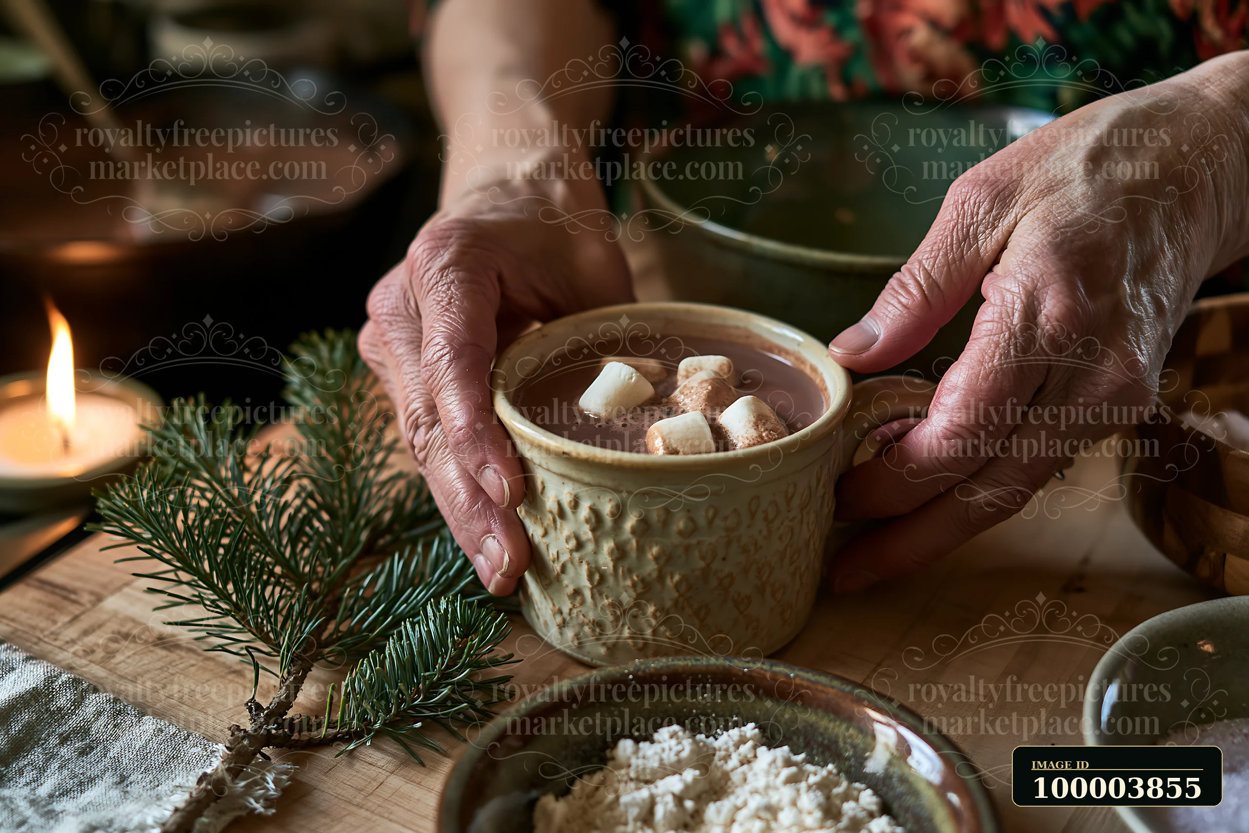 Hands hold a rustic mug of cocoa topped with marshmallows beside evergreen branches and a lit candle.
