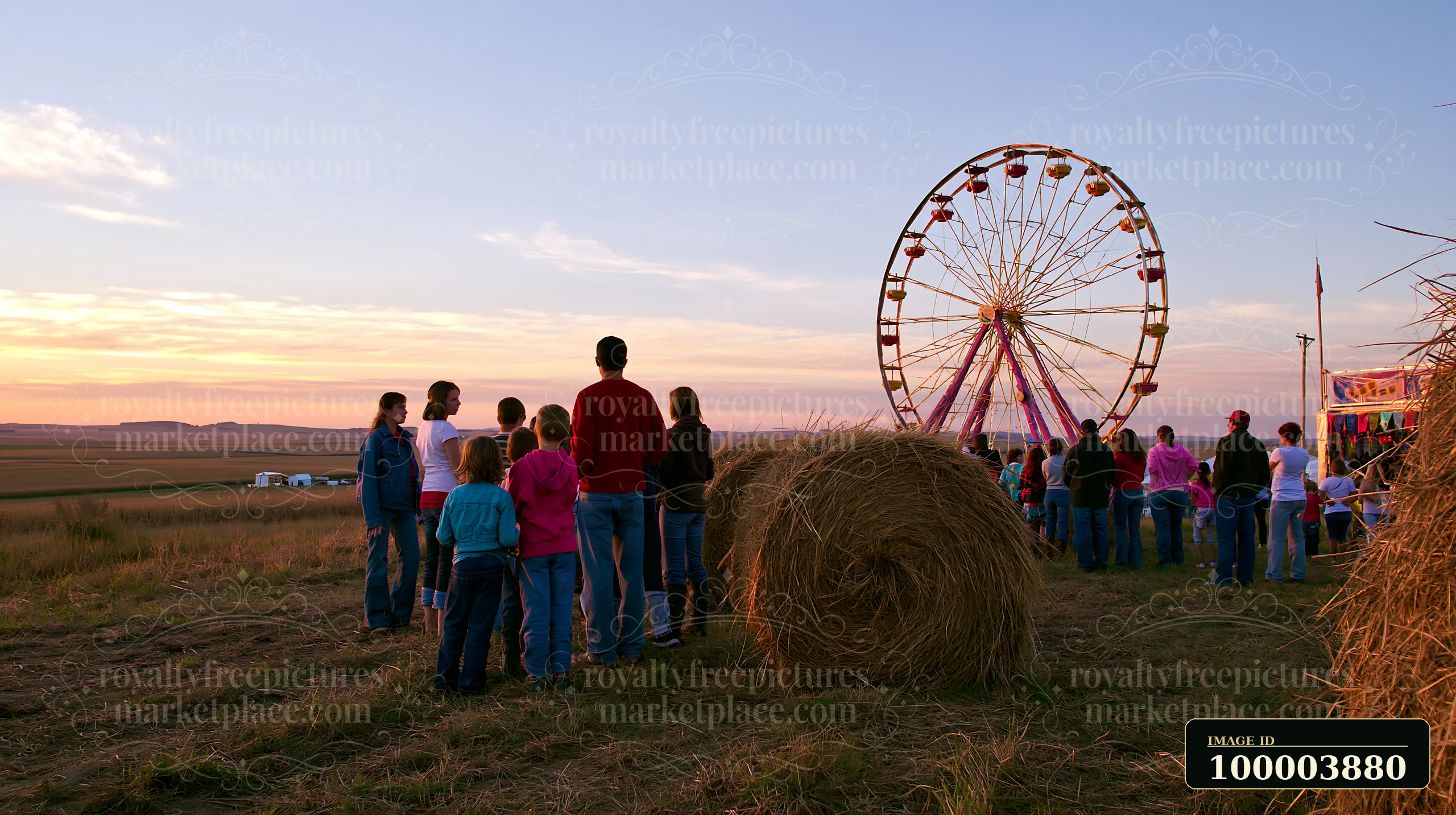 Families wait in line beside a hay bale for a Ferris wheel at an outdoor fair during sunset.
