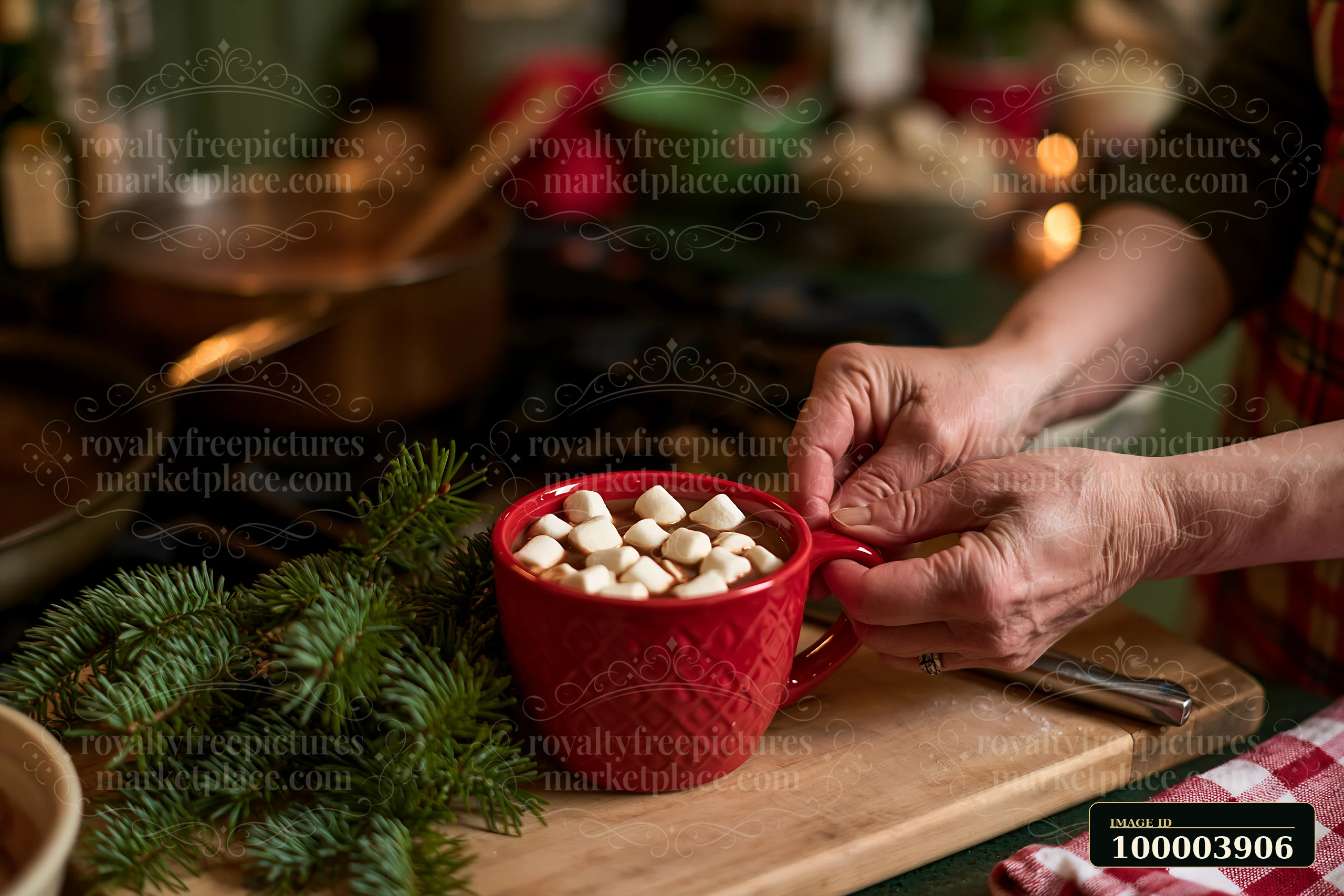 Hands hold a red mug of cocoa topped with marshmallows beside evergreen branches on a wooden board.