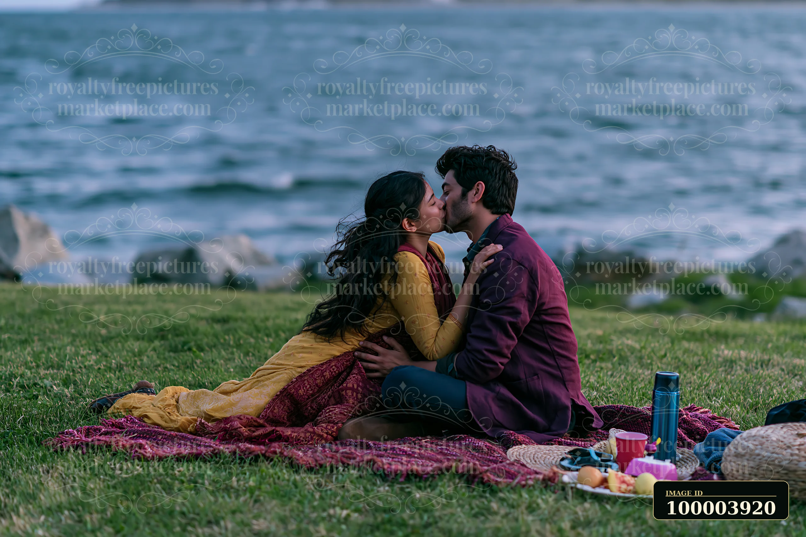 Couple kissing on a picnic blanket beside the sea with rocky shore and picnic items nearby.