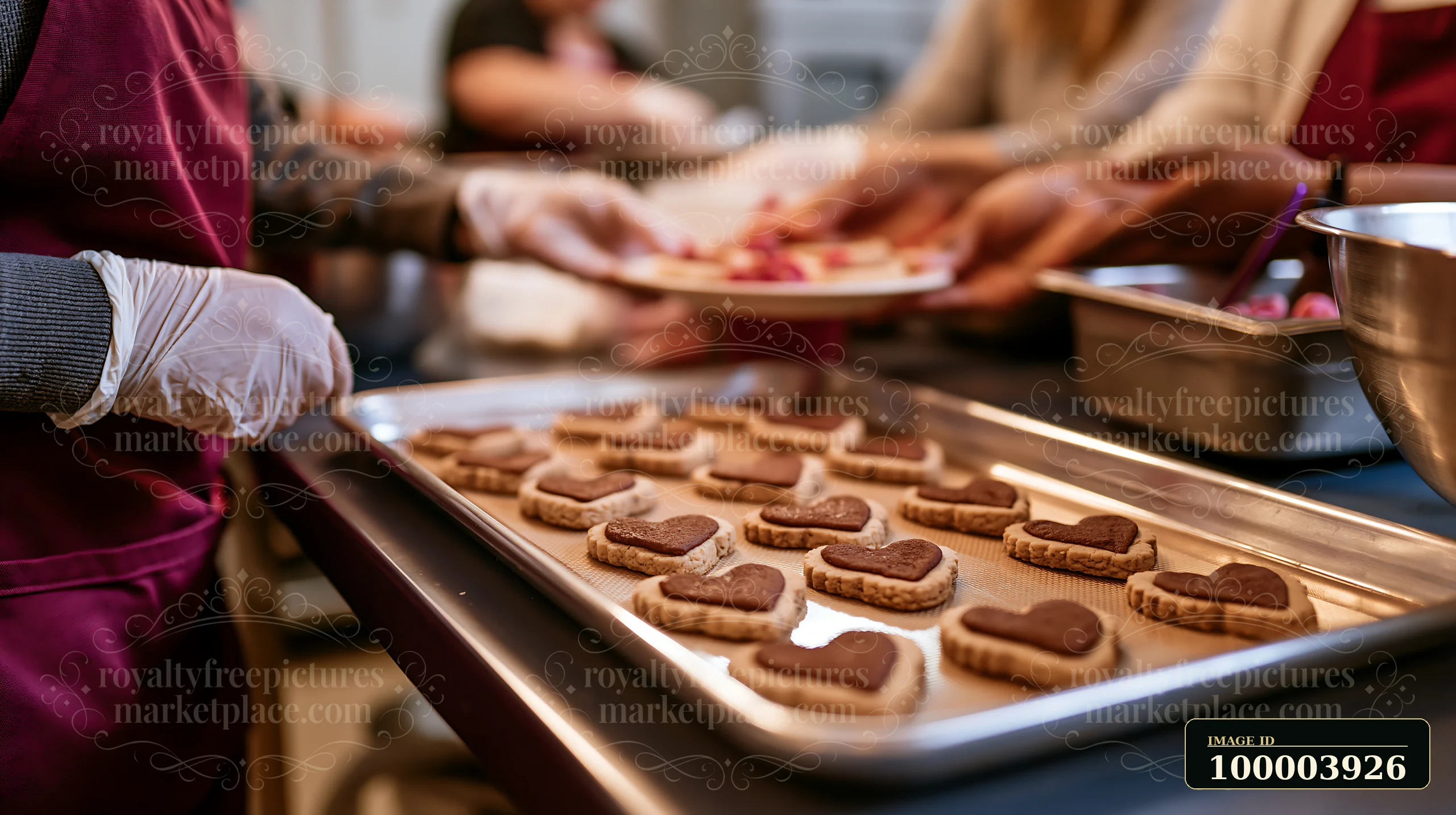 A person in gloves is decorating heart shaped cookies on a kitchen counter while others work in the background.