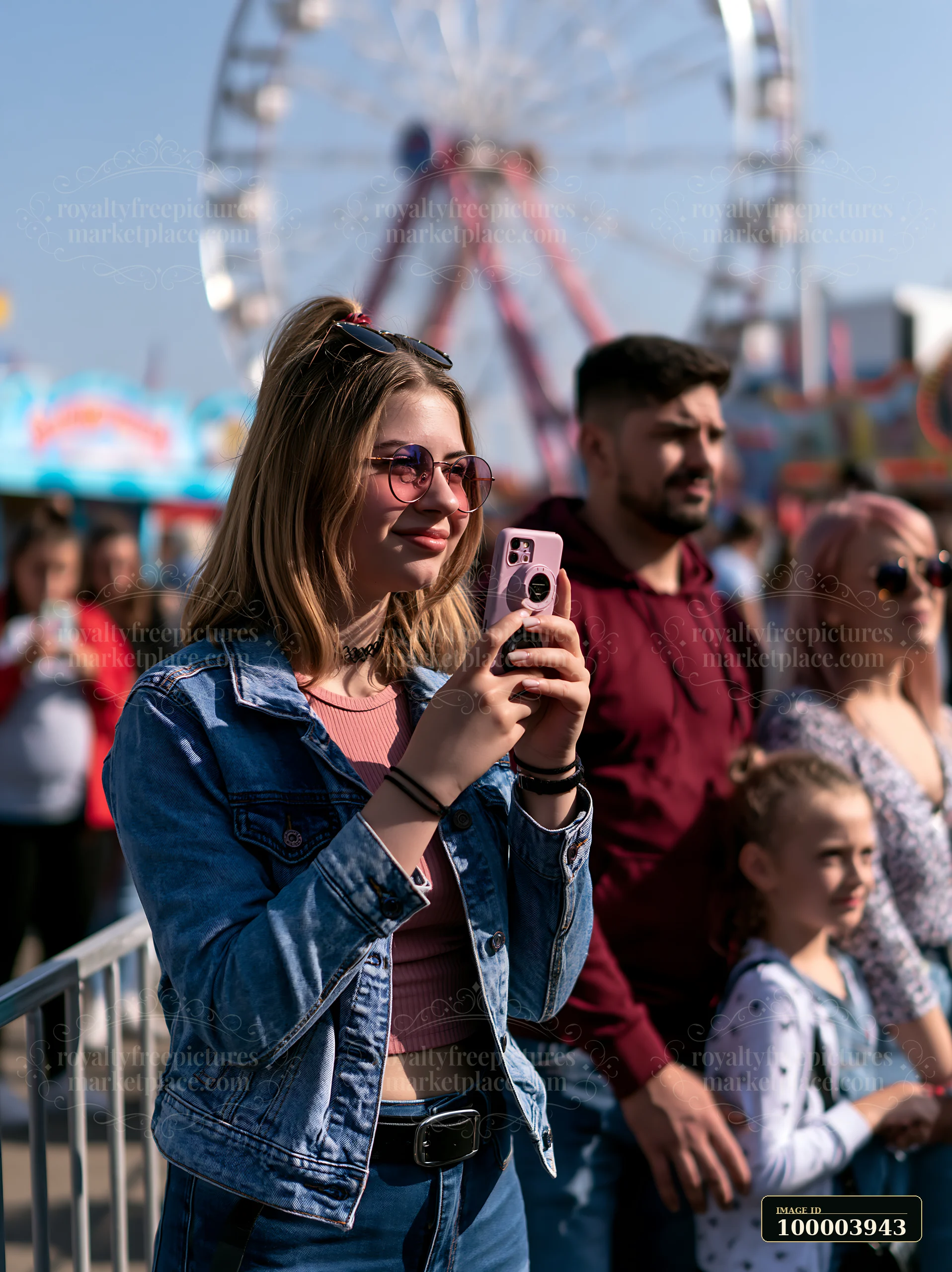 Family waits in a fairground queue while a young woman uses her smartphone near a blurred ferris wheel.