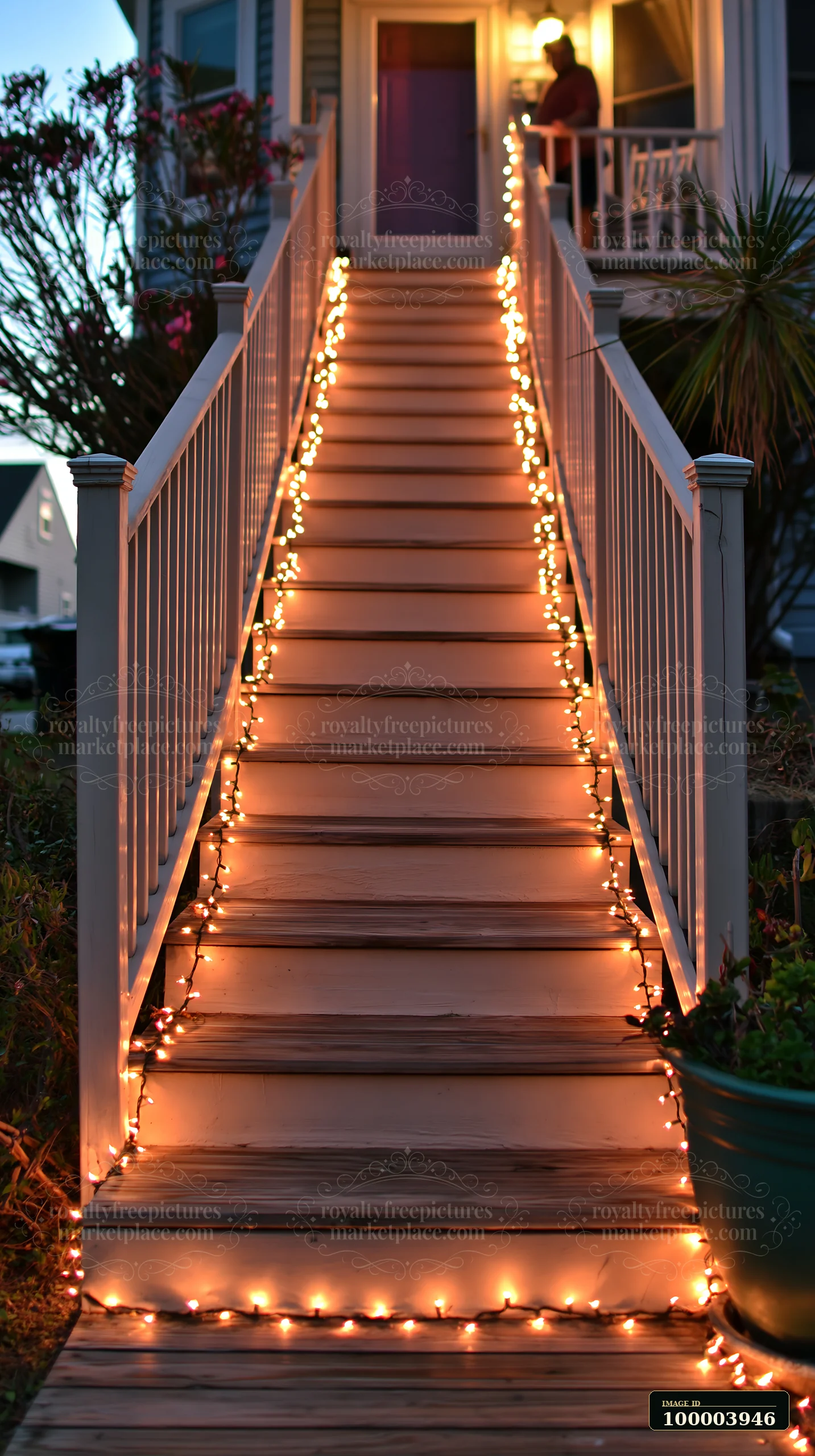Person hangs warm string lights along a wooden staircase outside a home at dusk.