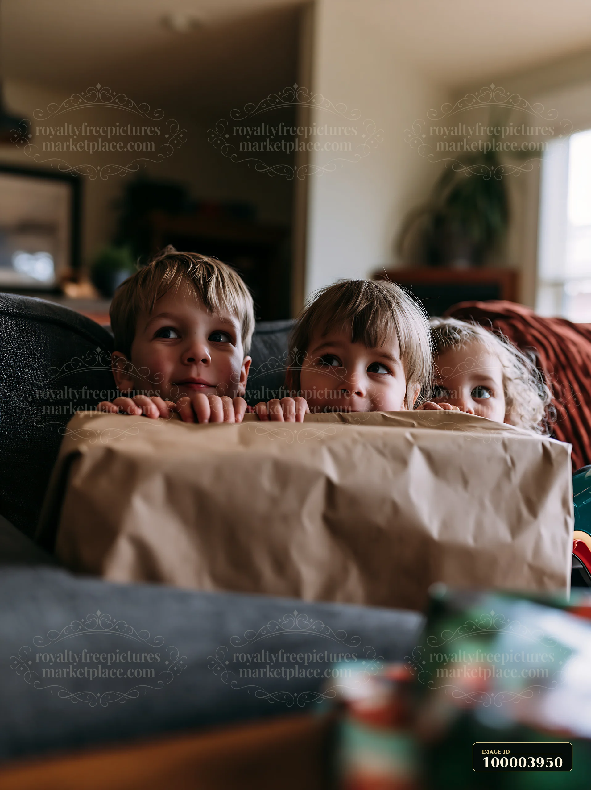 Three children peek over a large wrapped present in a warmly lit living room.