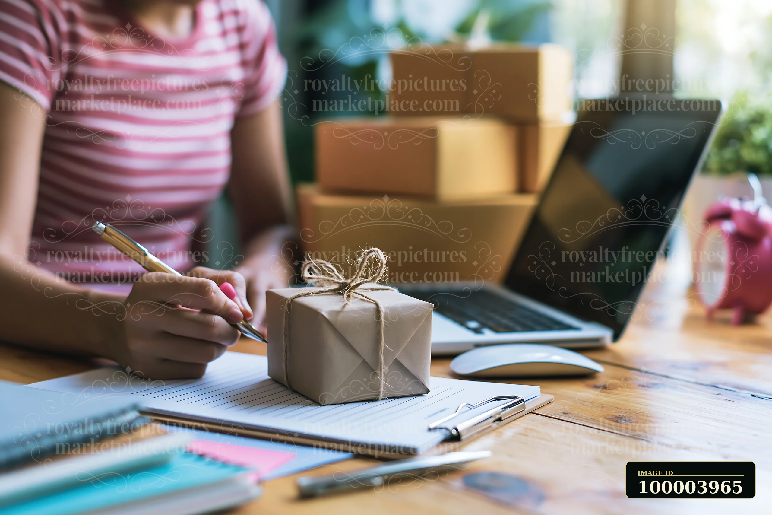 A person is writing a gift list at a desk with a laptop open and packages behind them.