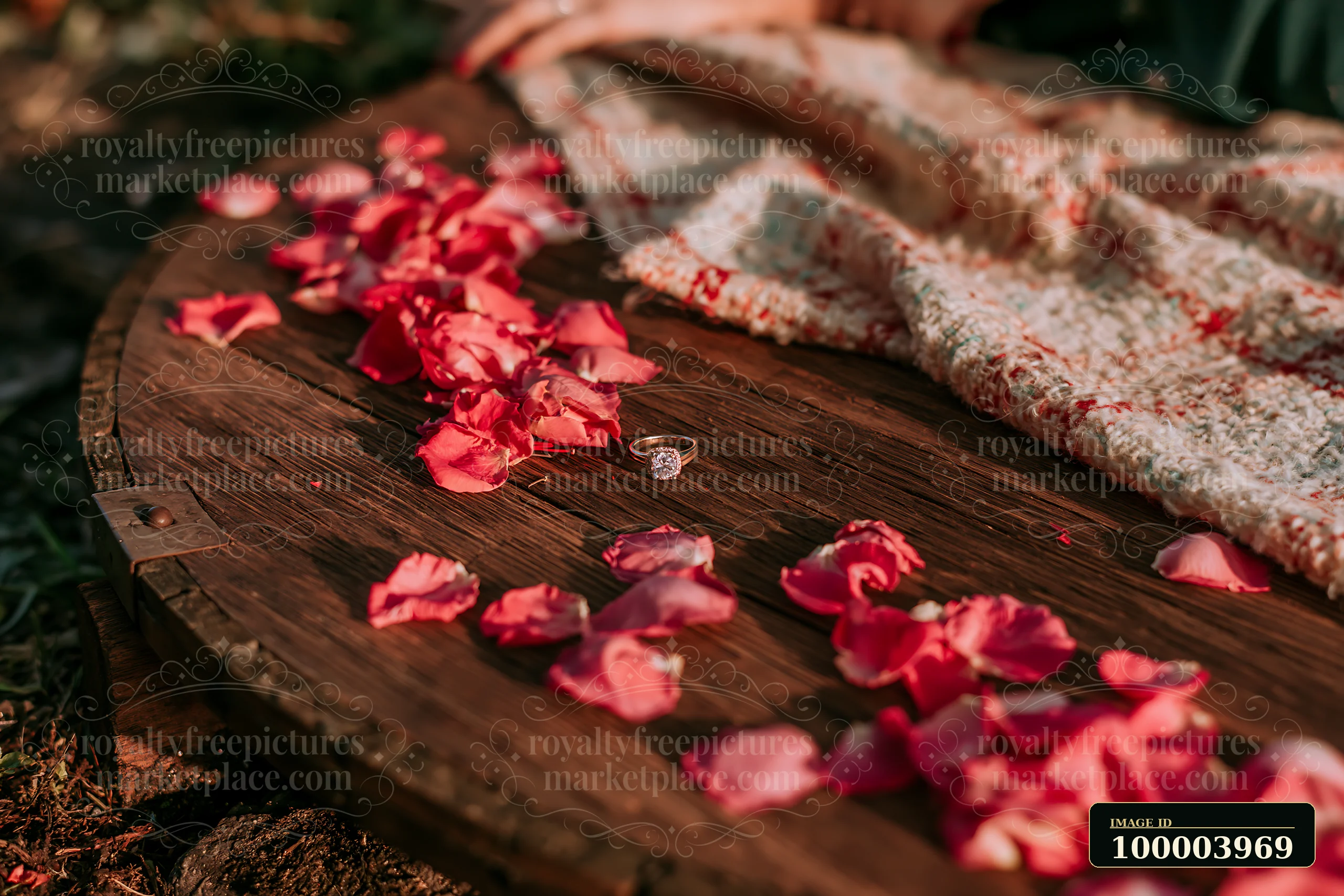 Engagement ring beside scattered rose petals on a rustic wooden table with a folded blanket.