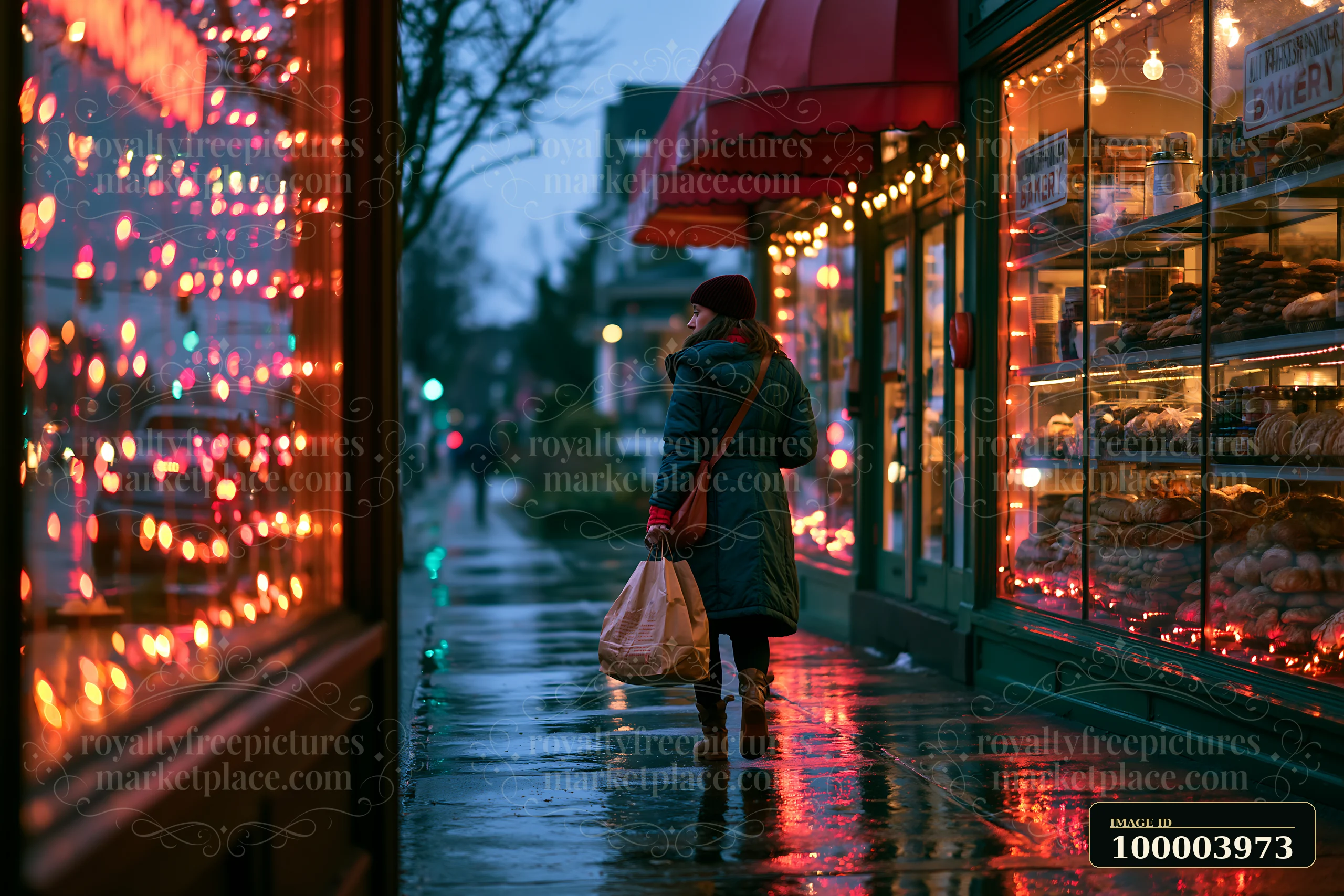 Woman carrying shopping bags walks past a warmly lit bakery on a wet street at dusk.