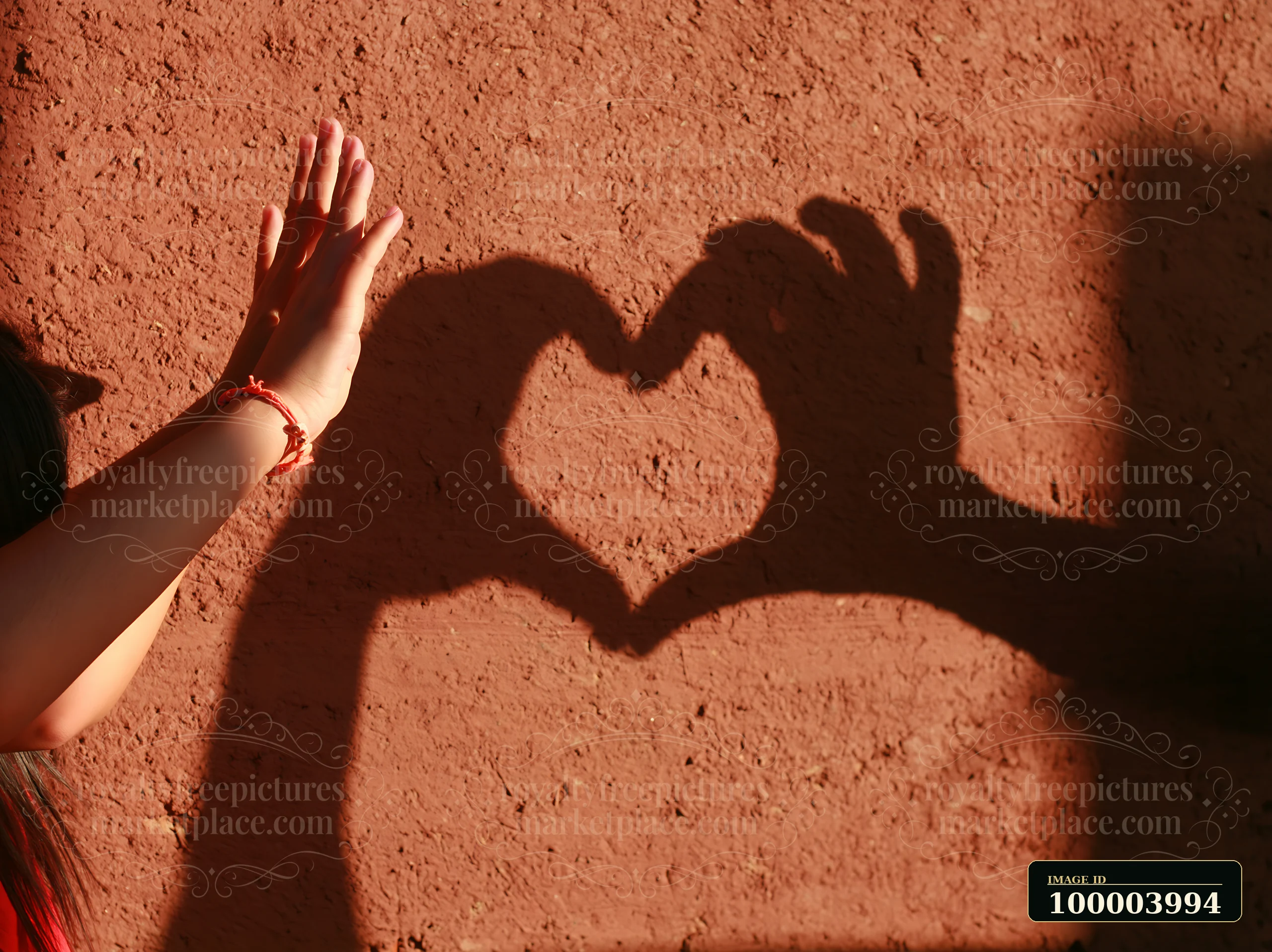 A hand reaches toward a heart-shaped shadow on a sunlit textured wall.