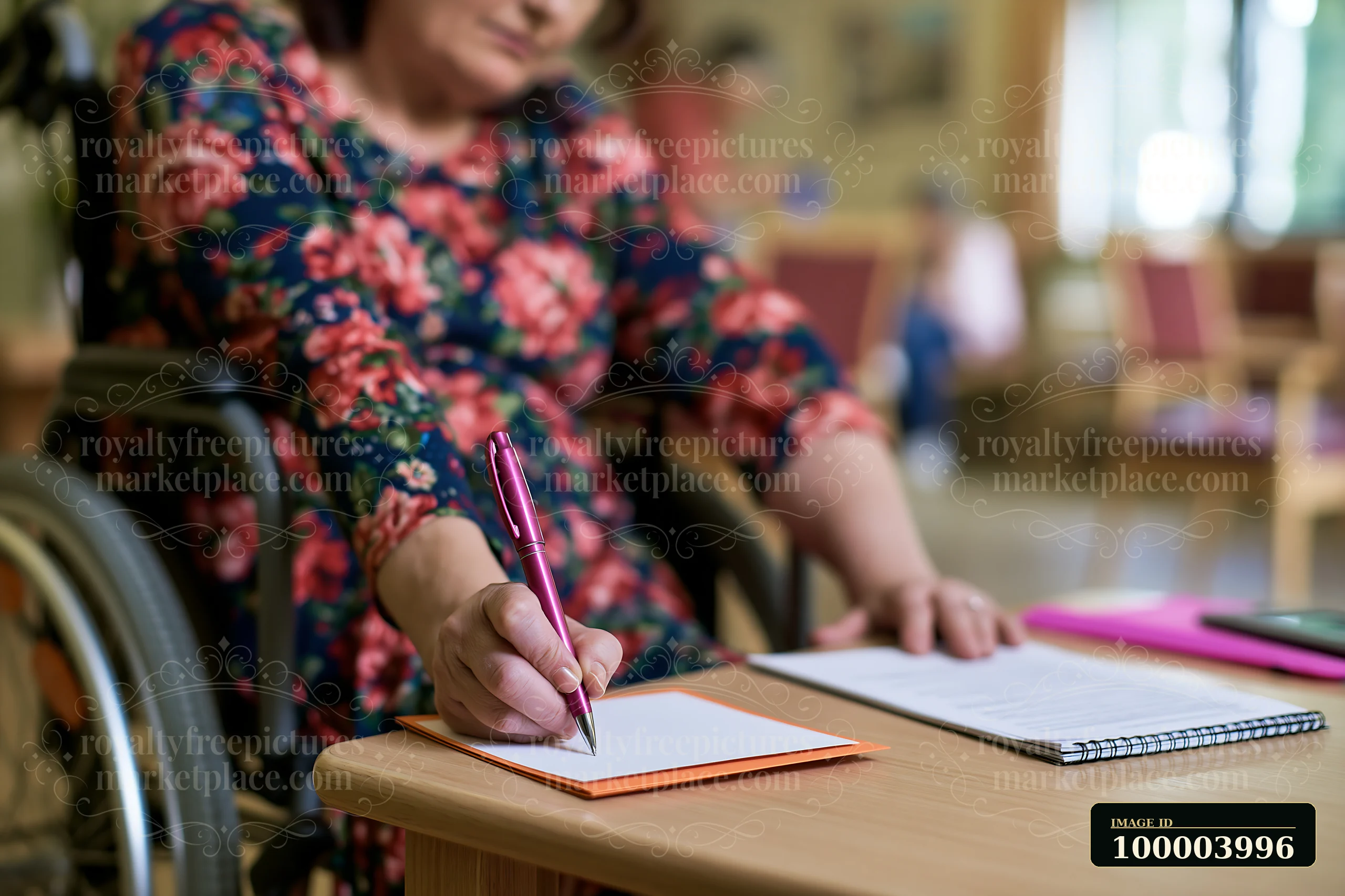 A woman in a floral dress is seated in a wheelchair, writing Christmas cards at a table in a care facility.