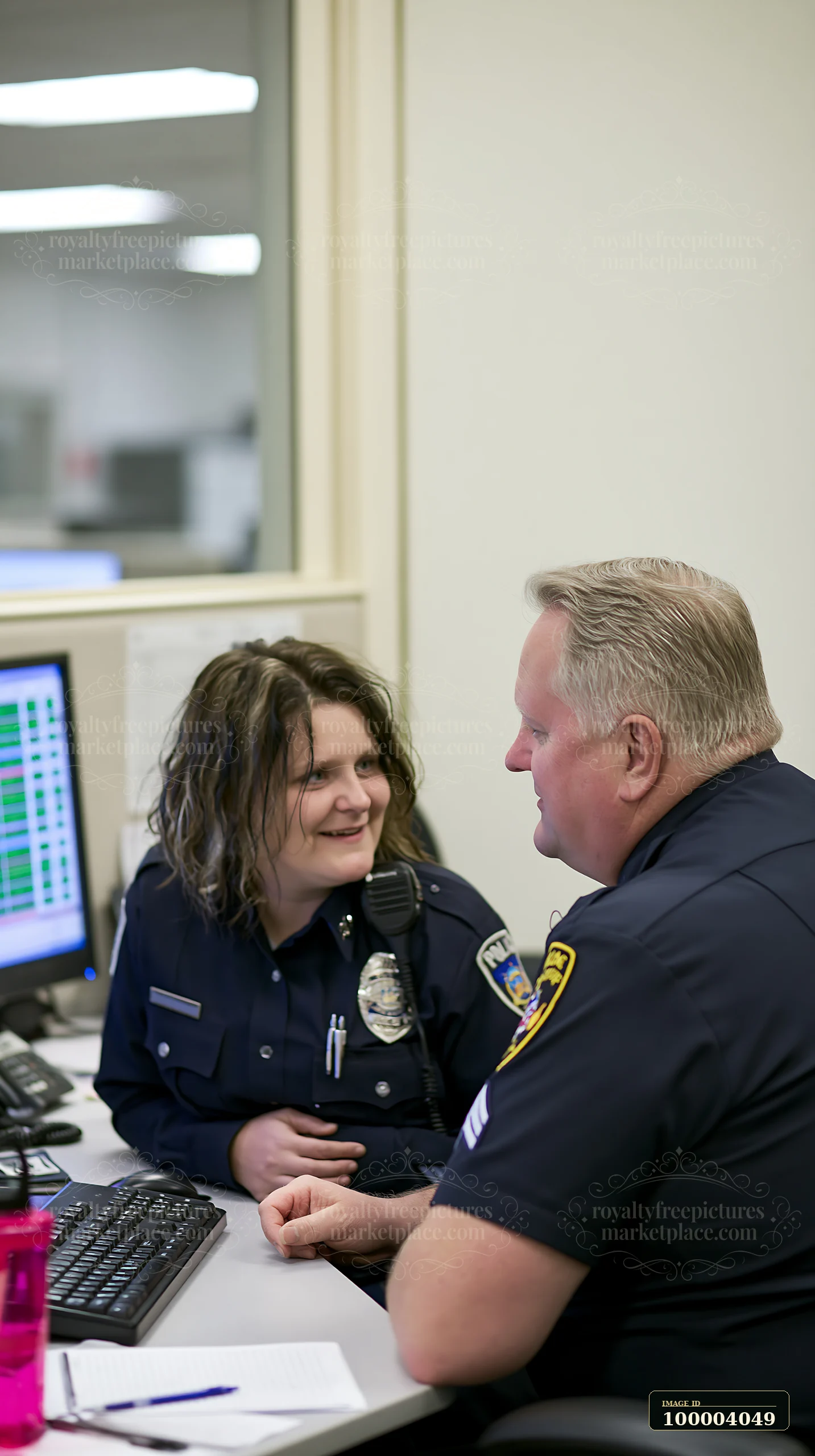 A police chief is talking with an officer at a desk in an office.