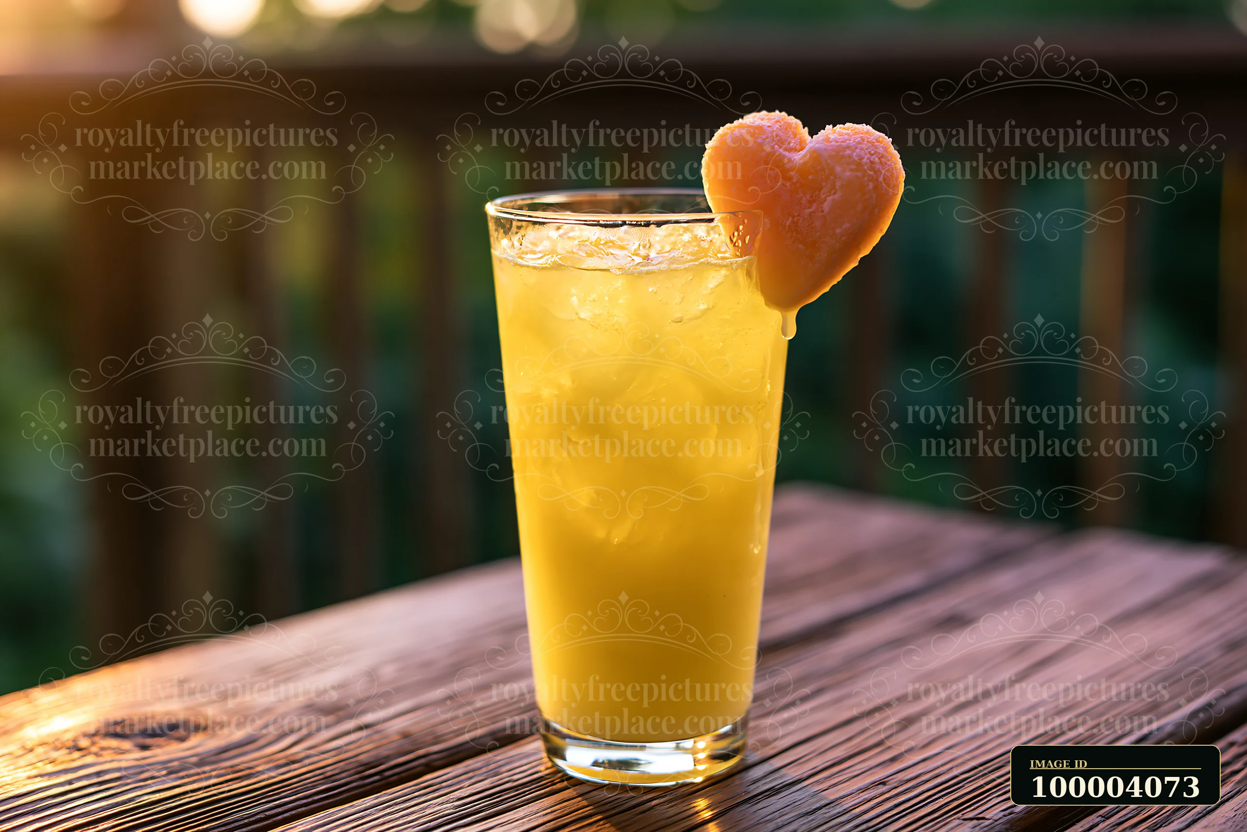 A heart-shaped ice cube melts in a glass of yellow drink placed on a wooden table outdoors.
