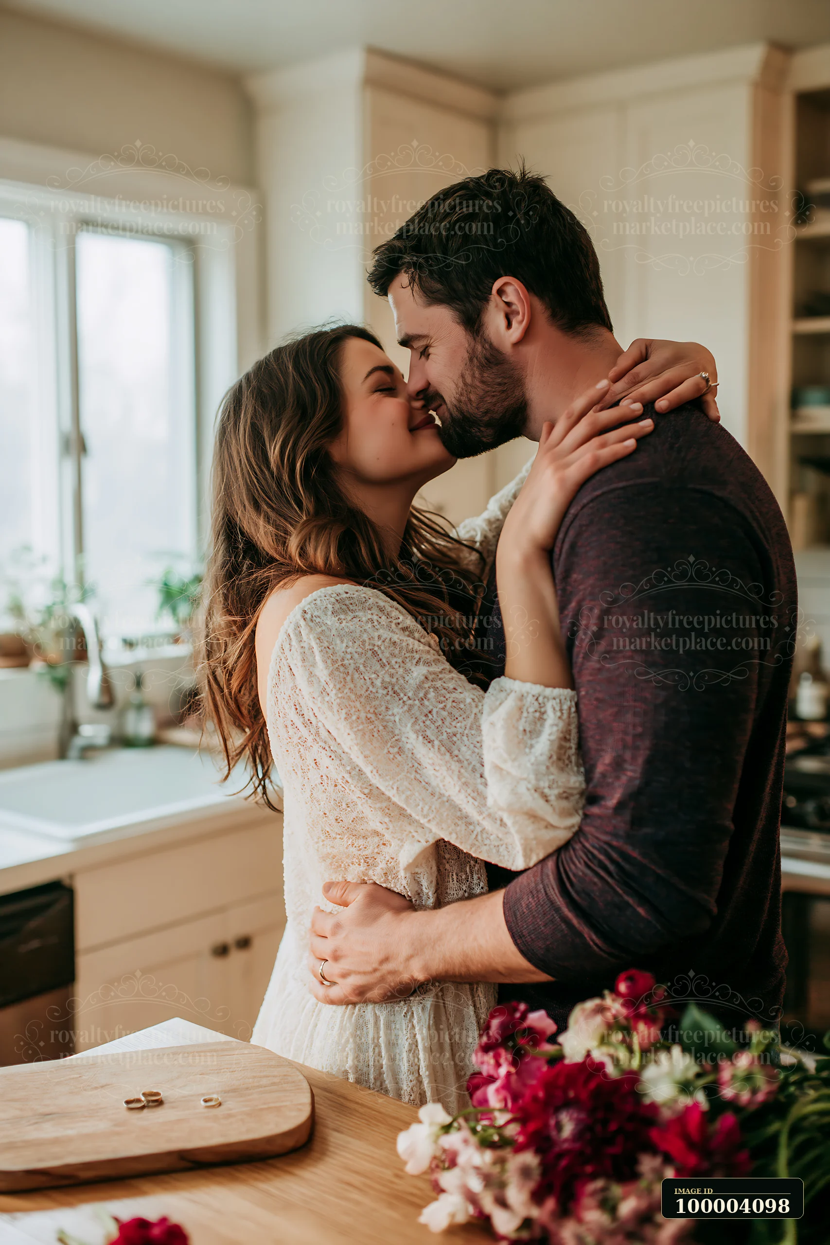 Couple kisses and embraces beside wedding rings on a wooden table in a warm kitchen.