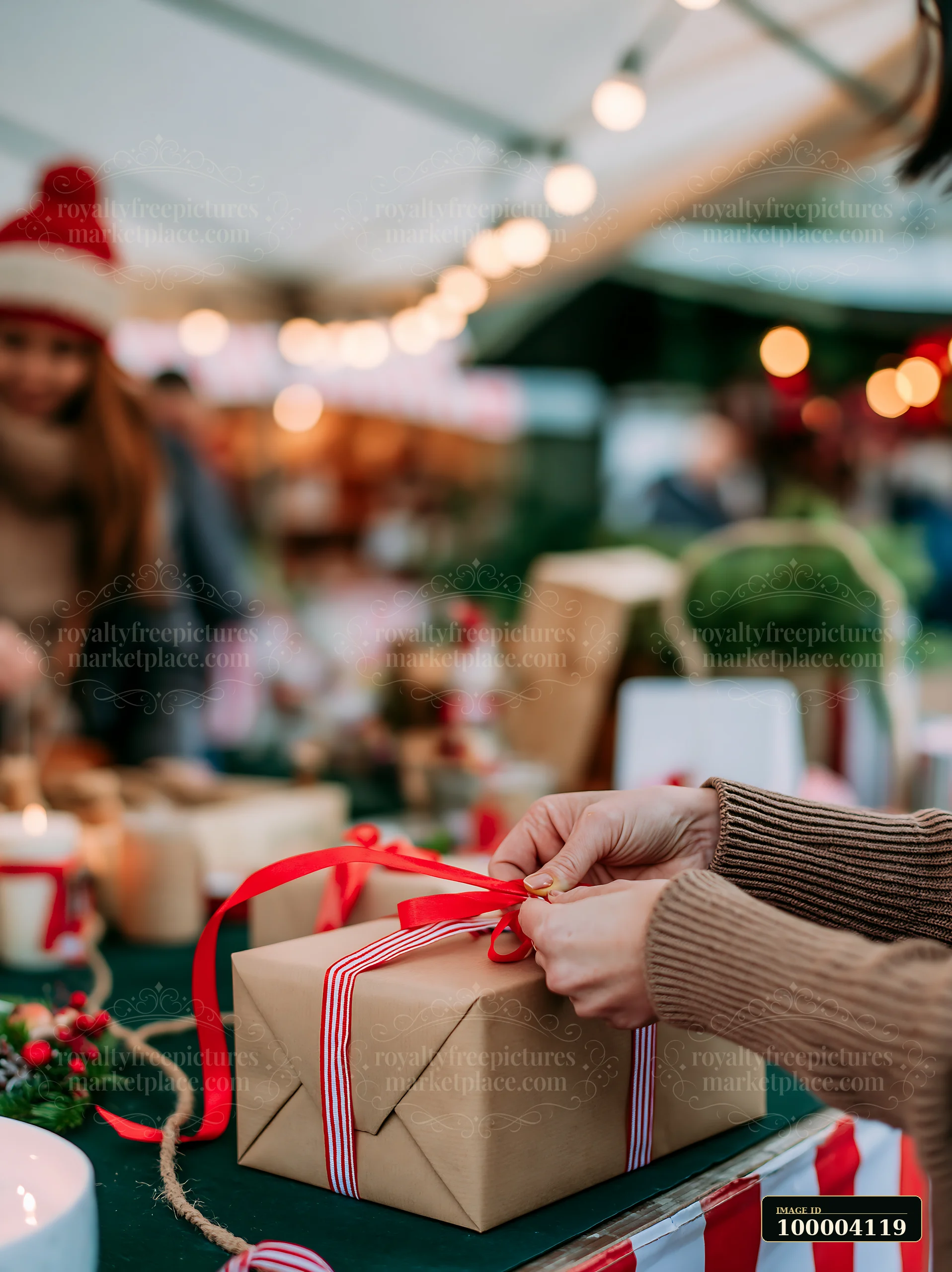 Hands are tying a red ribbon around a gift box while people shop at an outdoor market.