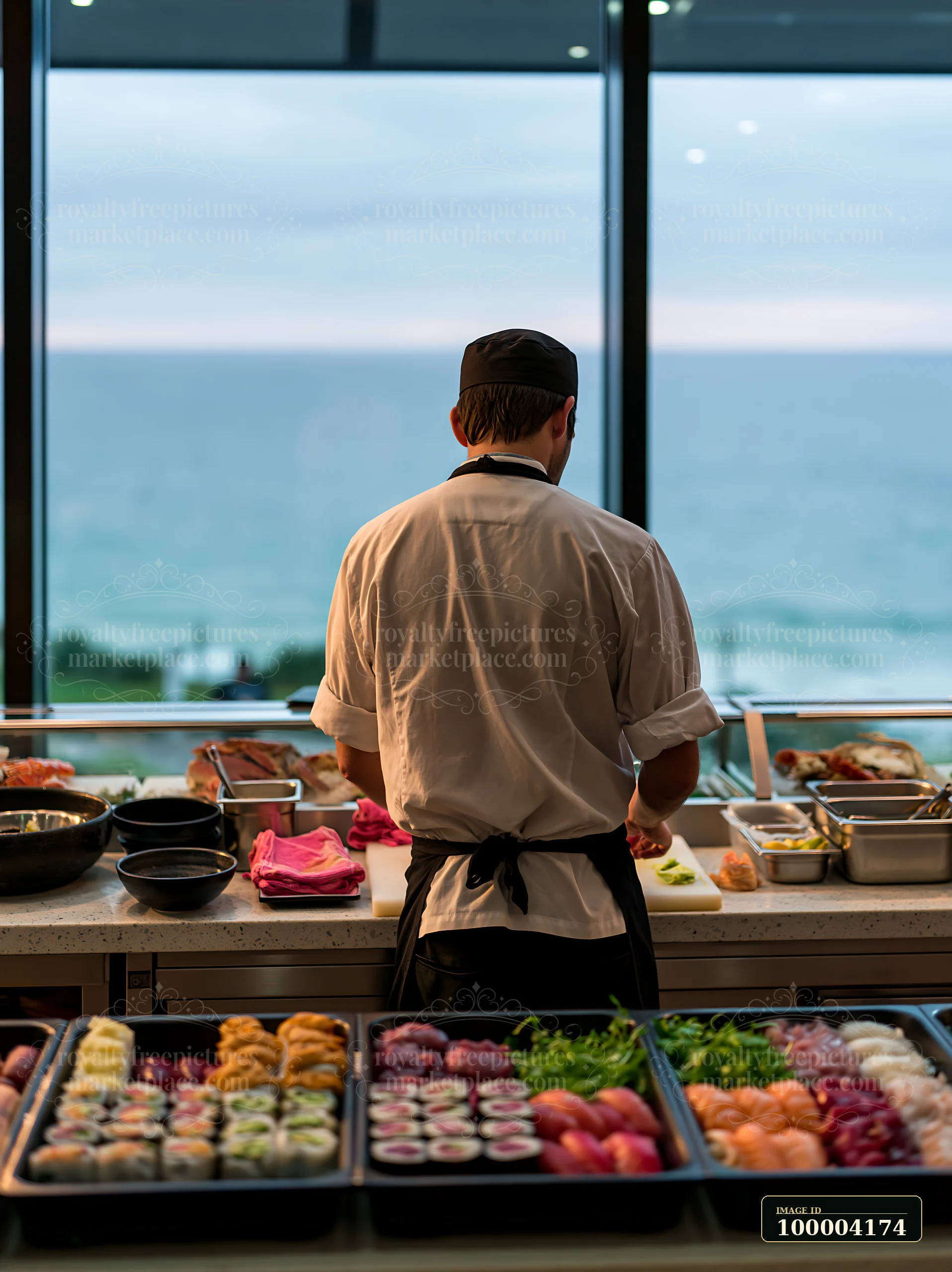 Sushi chef seen from behind at a preparation counter with sushi trays and a sea view.